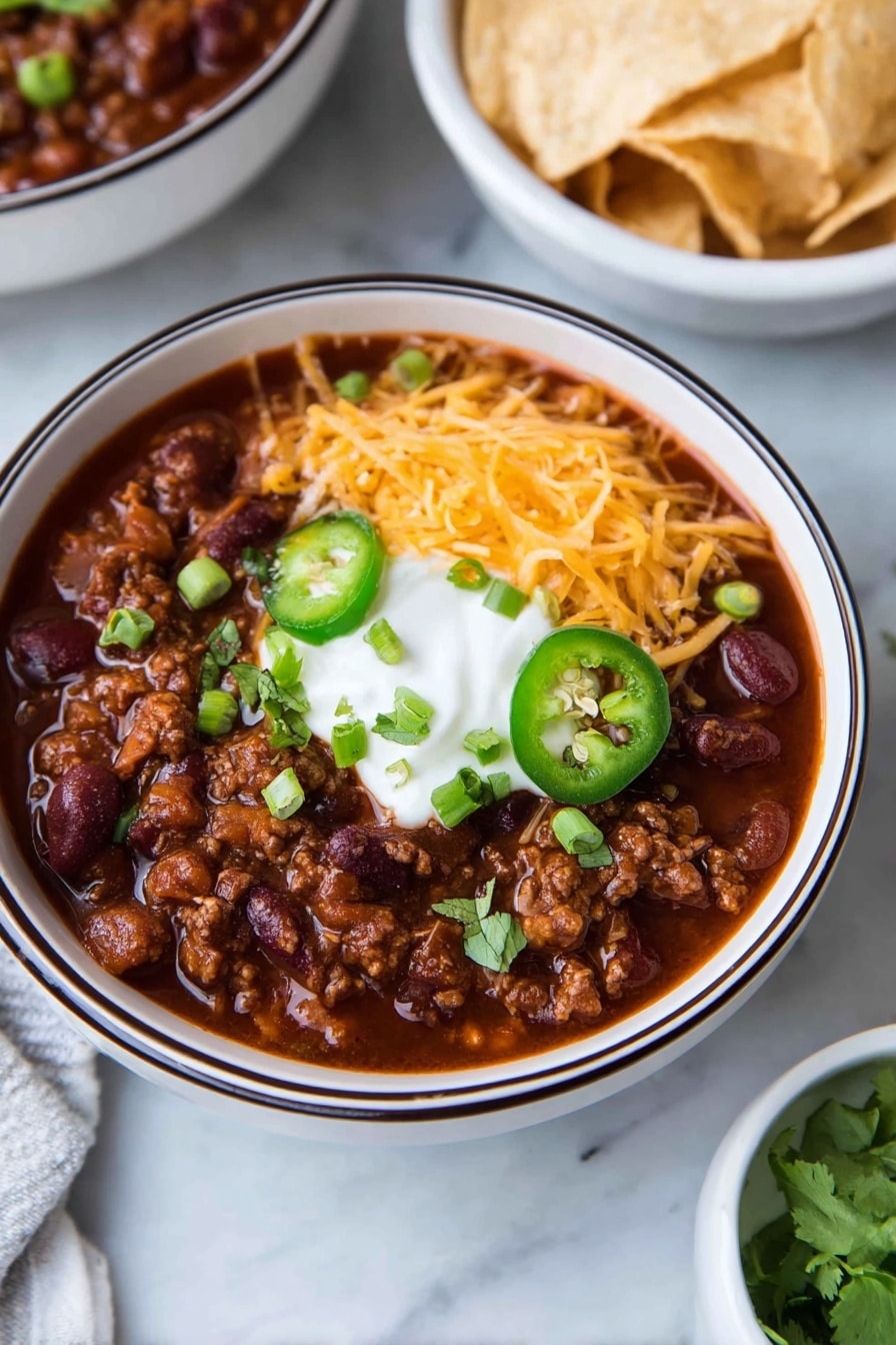 A white bowl with a dark rim holds a thick dark red chili filled with beans and ground meat. On top, there is a layer of shredded orange cheddar cheese on one side, a dollop of white sour cream in the center, sliced green jalapeño rings below, and chopped green onions and cilantro scattered around. In the background, there is a white bowl filled with light-colored tortilla chips on a white marbled surface. Photo taken with an iphone --ar 2:3 --v 7 - Hearty Turkey Chili, turkey chili recipe, healthy turkey chili, easy turkey chili, comforting chili with beans