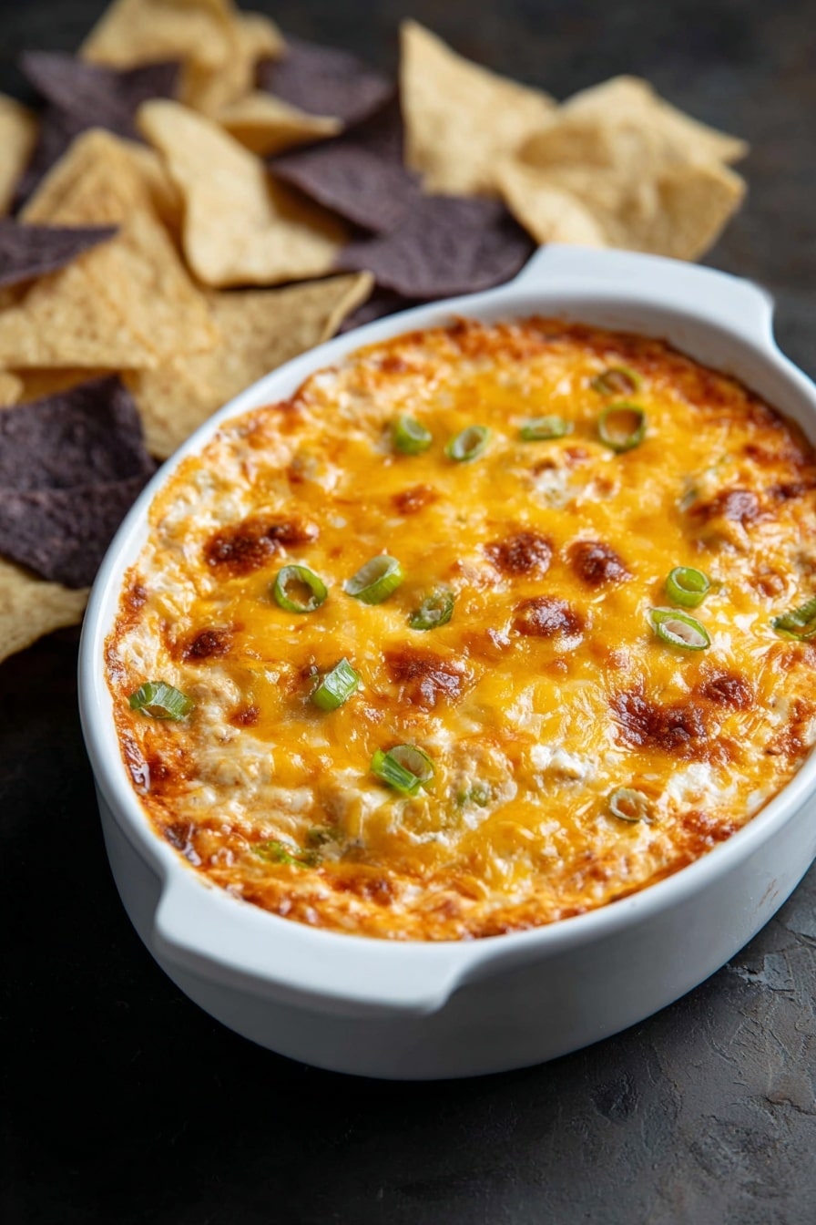The image shows a white oval dish filled with a baked dip that has a golden-brown melted cheese layer on top with some browned spots. The cheese surface is dotted with small green onion pieces, giving a pop of green color. The dip looks creamy with slightly uneven texture, suggesting layers of ingredients underneath the cheese. The dish sits on a dark surface, and there are light and dark tortilla chips placed around the dish in the background. photo taken with an iphone --ar 2:3 --v 7 - Buffalo Chicken Dip, spicy chicken dip, easy party appetizers, creamy buffalo dip, game day appetizer