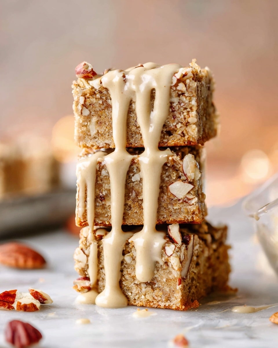 The image shows a stack of three square cake pieces placed vertically on a white marbled surface, each piece with a light brown nutty cake base filled with visible nuts and a creamy, beige frosting layer on top sprinkled with chopped pecans. Around the stack are scattered pecan pieces and red autumn leaves, adding warm colors to the scene. In the background, there are blurred brown pine cones, white candles, and a brass candle holder that create a cozy, soft atmosphere. A metal cake server lies on the surface at the front left, slightly out of focus. photo taken with an iphone --ar 4:5 --v 7