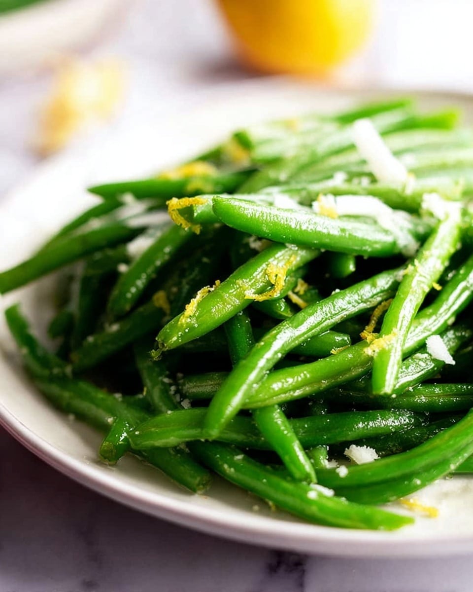 A white plate filled with bright green cooked green beans arranged in a slightly piled way, shiny with a light coating probably from oil or butter. Small bits of grated white ingredient, likely cheese or garlic, are scattered on top and some tiny specks of yellow zest add a fresh look. The background is a white marbled texture with a blurry out-of-focus yellow object in the far background. Photo taken with an iphone --ar 4:5 --v 7