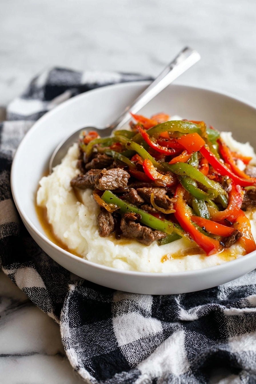 A white bowl holds a dish with two main layers: the bottom layer is a smooth, creamy white mashed texture, slightly spread around the edges. On top, there is a colorful mix of thin strips of cooked green, red, and orange bell peppers, mixed with small pieces of brown cooked meat, all glistening with a light sauce. A silver spoon rests inside the bowl, angled towards the back. The bowl sits on a black and white checkered cloth, placed on a white marbled surface. photo taken with an iphone --ar 2:3 --v 7 - Pepper Steak with Onion, Pepper Steak with Onion recipe, Easy Pepper Steak with Onion, Tender Pepper Steak with Onion, Beef and Onion Stir Fry