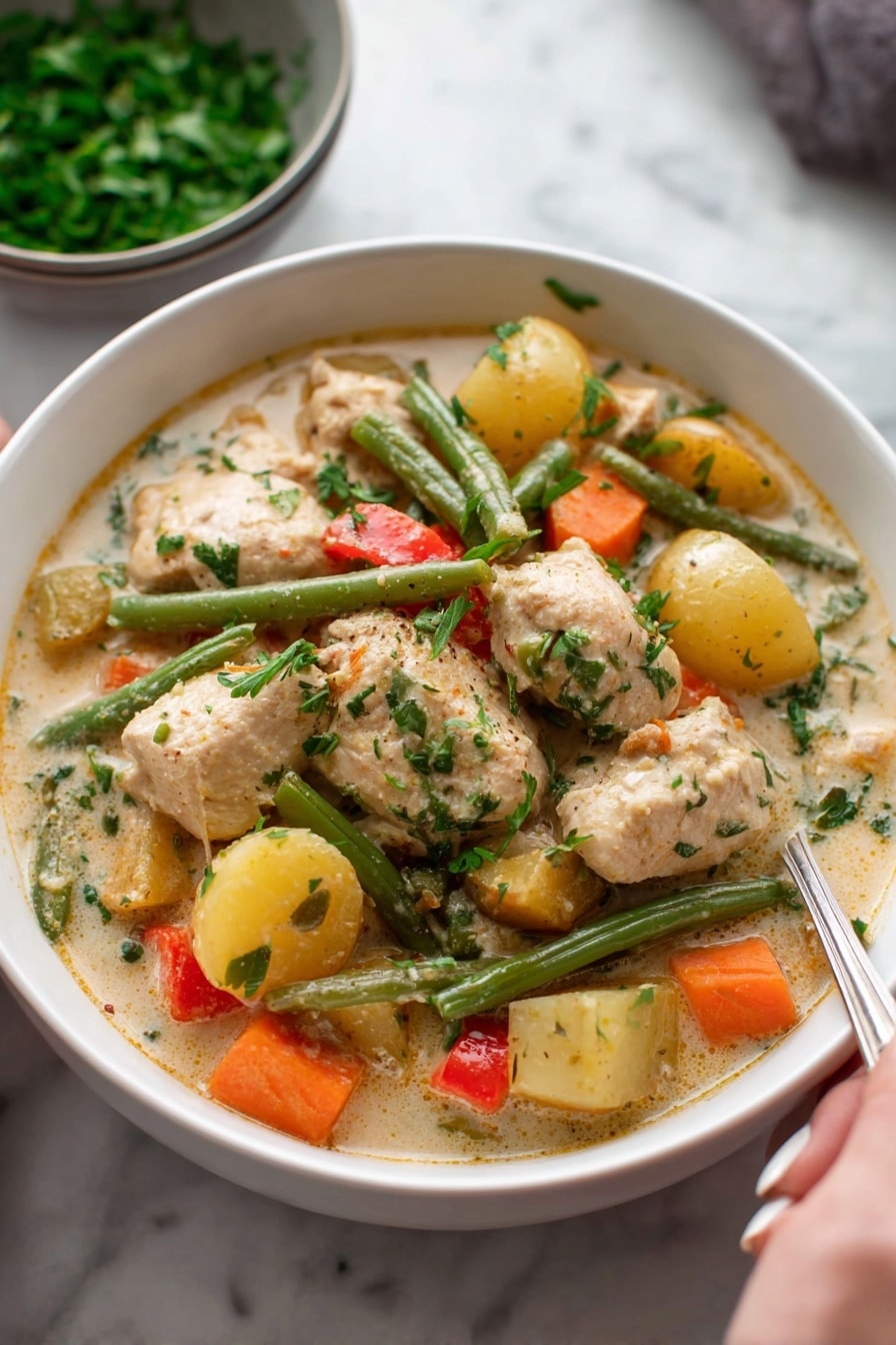 A white bowl filled with creamy chicken stew, showing large pieces of light brown chicken and chunky vegetables including orange carrots, green beans, yellow potatoes, and small bits of red pepper, all mixed in a thick, pale sauce with visible herbs on top; in the background, a bowl of chopped green herbs rests on a white marbled surface, and a woman's hand holding a spoon is partly visible at the bottom right. photo taken with an iphone --ar 2:3 --v 7 - Hearty Chicken Stew with Root Vegetables and Cream, chicken stew, root vegetable recipes, creamy chicken stew, comforting fall recipes
