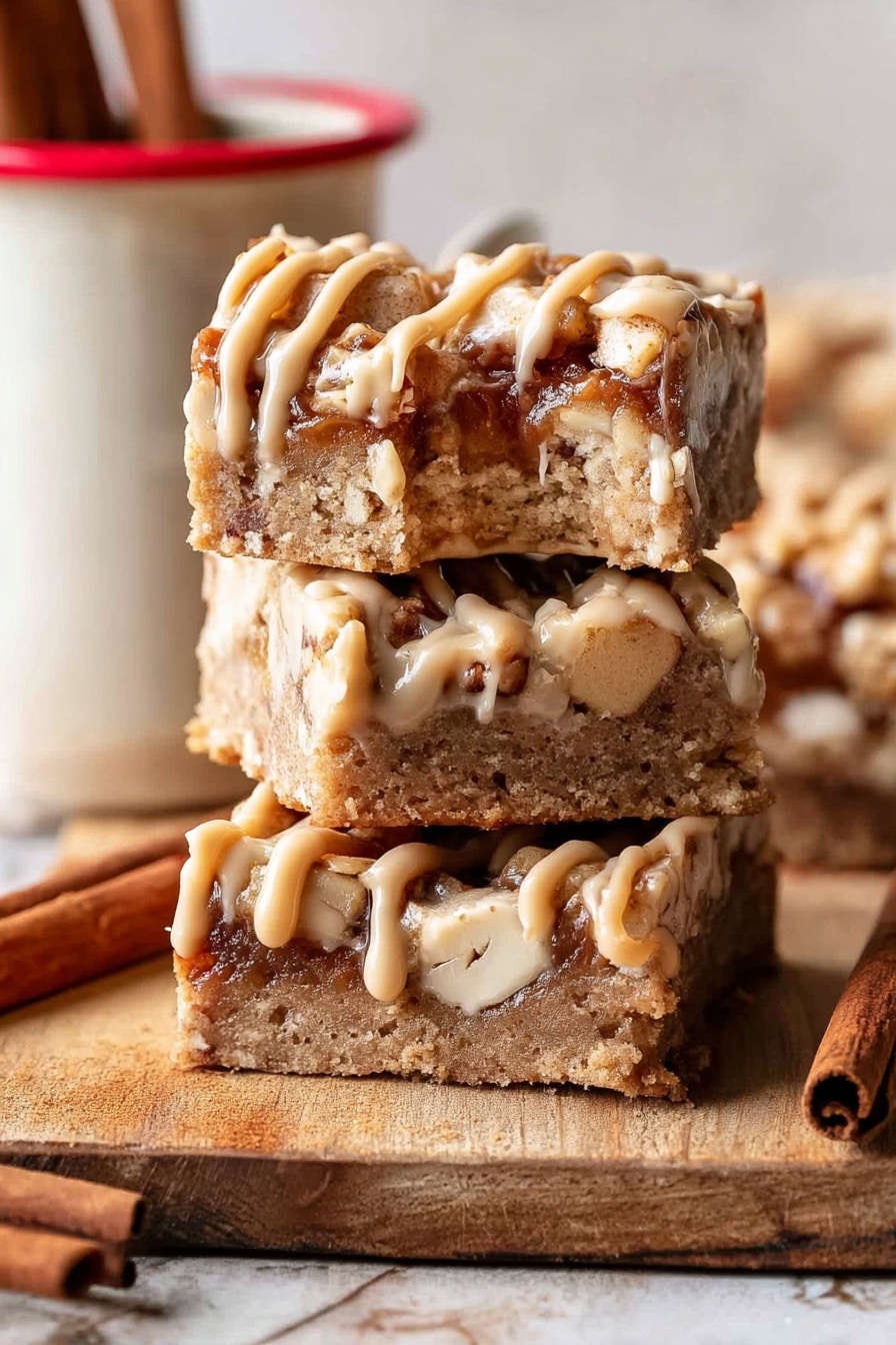 The image shows three square bars stacked vertically on a wooden surface with a single bite taken from the top bar. Each bar has a thick, crumbly base layer in light brown with small white nut pieces embedded inside. On top of the base, there is a layer of darker brown chunks with a soft texture, drizzled with thick, pale caramel-colored frosting in thin, uneven lines. A cinnamon stick is placed near the bottom left corner, and a white mug with a red rim and two cinnamon sticks inside is partially visible to the right. The background is a white marbled texture. Photo taken with an iphone --ar 2:3 --v 7 - Brown Butter Apple Blondies with Cinnamon Maple Glaze, fall apple blondies, cozy dessert recipes, cinnamon glazed blondies, easy apple dessert