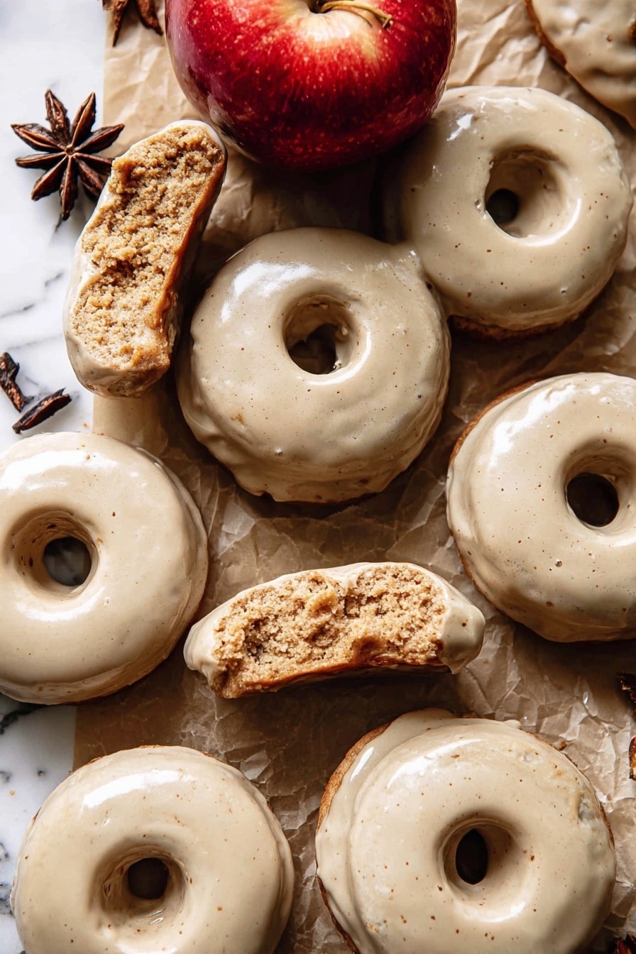 The image shows a group of doughnuts with a smooth, light beige glaze covering their top surfaces. Each doughnut has one round hole in the center and a slightly rough, crumbly texture inside where some are broken or bitten. The doughnuts are laid on a brown paper surface with pieces of white parchment paper scattered around. There is a shiny red apple near the center top of the image and a dark brown star anise near the upper left side. The background is a white marbled texture. photo taken with an iphone --ar 2:3 --v 7 - Baked Apple Cider Doughnuts with Cinnamon Maple Glaze, apple cider doughnut recipes, fall baked doughnuts, cinnamon maple glaze donuts, cozy autumn dessert