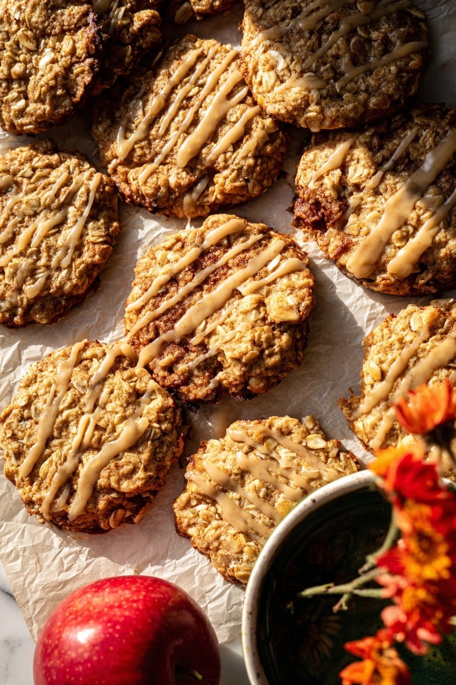The image shows a close-up of several oatmeal cookies with a slightly rough texture from oats and nuts, arranged unevenly on parchment paper over a white marbled surface. Some cookies are stacked, while others lay flat, revealing their golden-brown baked edges with a few darker spots. A few cookies are drizzled with a light beige glaze in thin, irregular lines, adding shine and contrast to the surface. A bright red apple is partially visible in the bottom left, and the corner of a white bowl with a dark liquid and a yellow flower is seen in the top right, adding color variety. The warm lighting highlights the cookies' texture and the glaze’s glossiness. photo taken with an iphone --ar 2:3 --v 7 - Iced Apple Cider Caramel Oatmeal Cookies, apple cider caramel cookies, oatmeal caramel cookies, fall cookies with caramel and oats, easy apple cider cookies