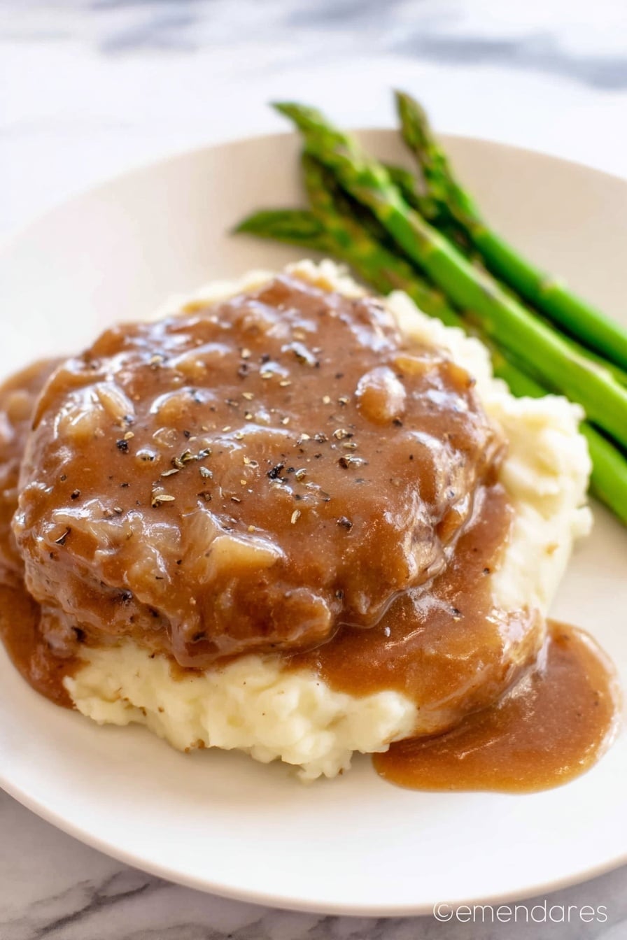 The image shows a white plate with three layers of food. The bottom layer is creamy mashed potatoes, which look soft and smooth in a pale cream color. On top of the mashed potatoes is a thick piece of cooked meat covered generously with a brown gravy sauce that has small onion bits and black pepper sprinkled on top. To the side of the meat, there is a small bunch of cooked green asparagus spears that add a fresh green color. The white plate sits on a white marbled surface. Photo taken with an iphone --ar 2:3 --v 7 - Crock Pot Cube Steak, slow cooker cube steak recipe, easy crock pot steak, tender cube steak slow cooker, comforting slow cooker dinner