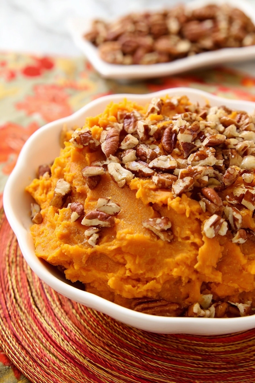 A white scalloped bowl holds a single layer of smooth, bright orange mashed sweet potatoes topped generously with a layer of chopped pecans, which add a textured, brown, and crunchy contrast. In the background, a white dish contains more pecan pieces, slightly out of focus. The bowl and pecans rest on a woven, multi-colored orange and red placemat, with a shiny silver spoon placed nearby. The scene is set on a white marbled surface. Photo taken with an iphone --ar 2:3 --v 7 - Slow Cooker Sweet Potato Casserole with Maple Syrup and Toasted Pecans, sweet potato casserole, holiday side dish, easy slow cooker sweet potato recipe, cozy fall casserole