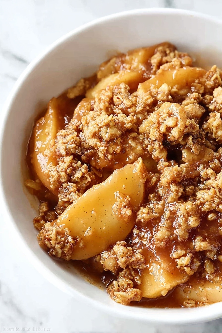A white bowl holds a warm apple dessert with three distinct layers: the bottom layer has soft, golden-brown apple slices coated in syrupy juices; above this is a crumbly, textured topping made of oats and brown sugar that looks crunchy and slightly toasted; the top layer is a smooth, creamy white scoop of melted vanilla ice cream that softly spreads over the crumble. A silver spoon rests in the bowl, partially visible at the back, all placed on a white marbled surface. Photo taken with an iphone --ar 2:3 --v 7 - Healthy Apple Crisp with Oat Pecan Topping, healthy apple crisp recipes, nutritious apple crisp dessert, wholesome apple crisp dessert, easy healthy apple crisp