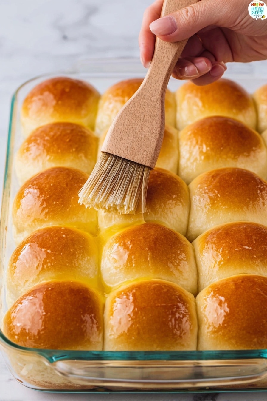 A glass baking dish holds a layer of 12 evenly arranged soft dinner rolls, each roll golden brown on top and shiny from being brushed with melted butter. The rolls sit closely together in a grid pattern with a soft, slightly fluffy texture. A woman's hand holds a light wooden brush with brown bristles, applying butter to the top of one roll. The background is a white marbled surface. photo taken with an iphone --ar 2:3 --v 7 - Homemade Fluffy Dinner Rolls, soft dinner rolls, buttery dinner rolls, easy dinner rolls recipe, homemade bread rolls