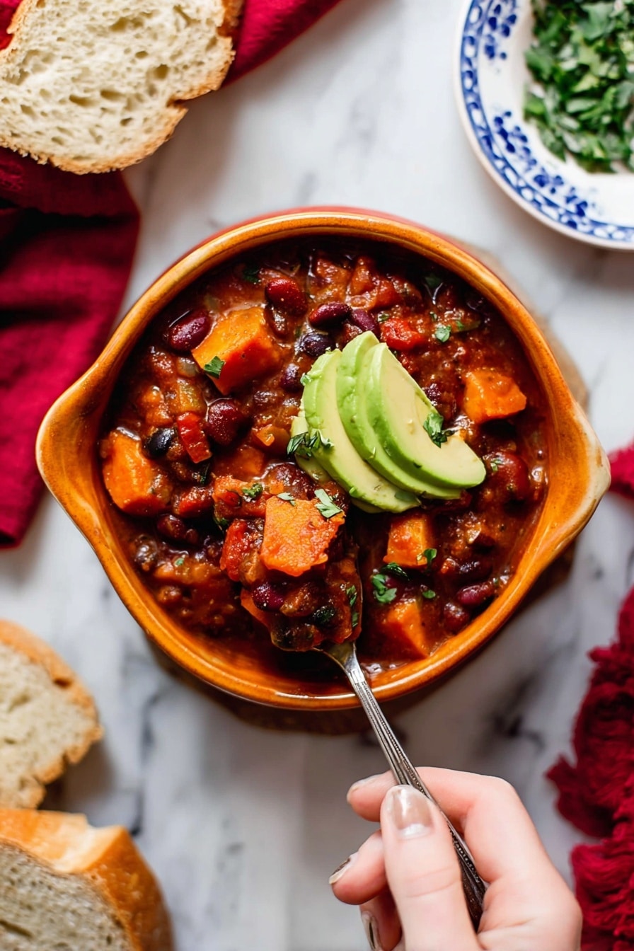 A bowl of thick chili with visible layers of chunky tomato sauce, red and black beans, and soft, cubed orange sweet potatoes, topped with two slices of green avocado. The chili is served in a curved, orange bowl on a white marbled surface, with a woman's hand holding a spoon scooping into the chili. Surrounding the bowl are slices of bread on a white plate with blue patterns, a small bowl of chopped green herbs, and a red cloth in the background. Photo taken with an iphone --ar 2:3 --v 7 - Vegetarian Pumpkin Chili, Best Pumpkin Chili, Easy Vegetarian Chili, Hearty Pumpkin Chili Recipe, Cozy Fall Chili