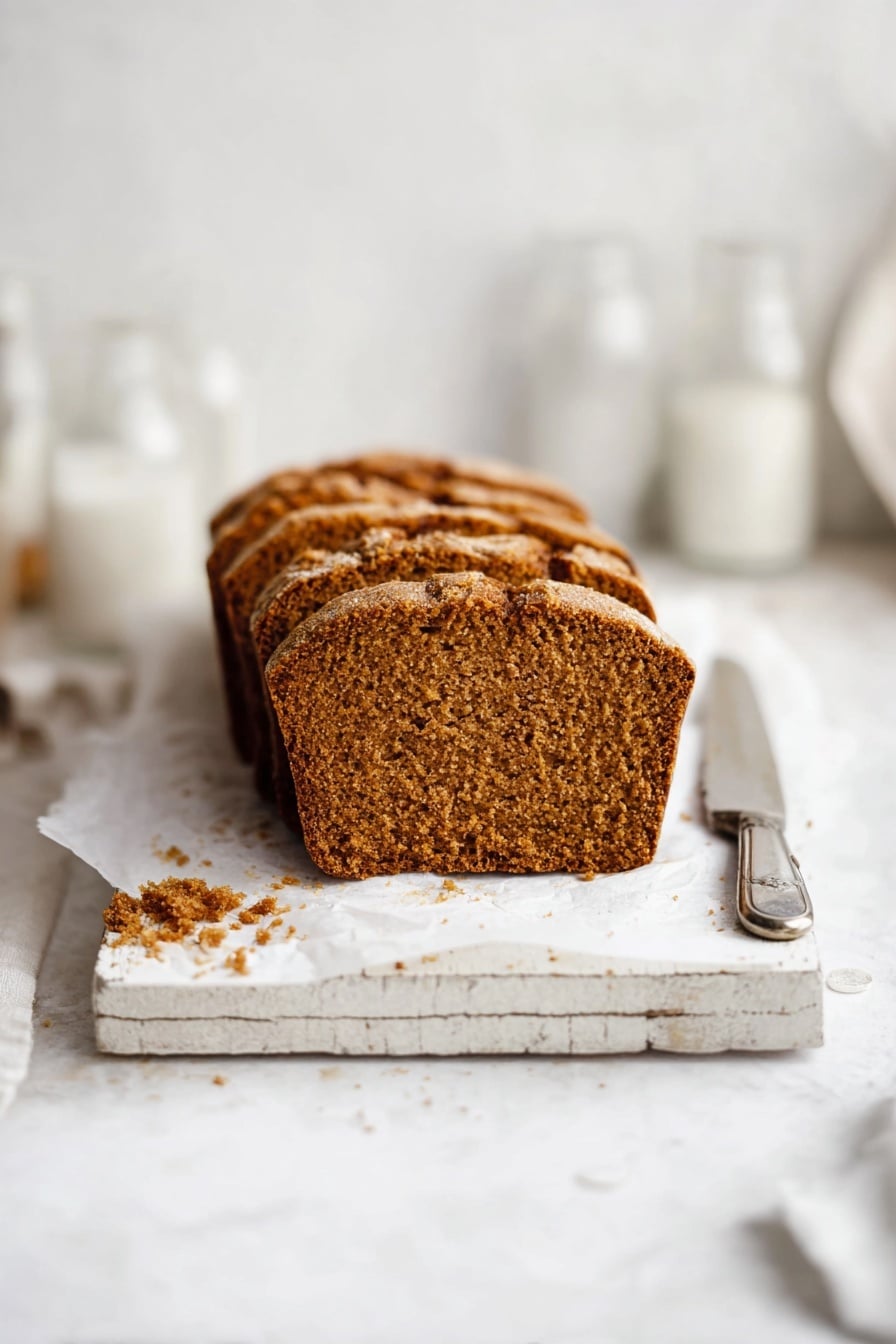 The image shows a loaf of brown bread sliced into six pieces, placed on white parchment paper on a rustic white wooden board. The bread has a golden-brown crust with a slightly rough texture and a dense, soft interior with small air holes. There are some crumbs scattered around the bread on the white marbled surface. To the upper right of the bread, there is a silver knife resting on top of a folded piece of white paper with text, and in the upper left, a clear empty glass and a white container are partially visible. Photo taken with an iphone --ar 2:3 --v 7 - Moist Pumpkin Bread, pumpkin bread recipe, fall pumpkin bread, easy pumpkin bread, tender pumpkin loaf