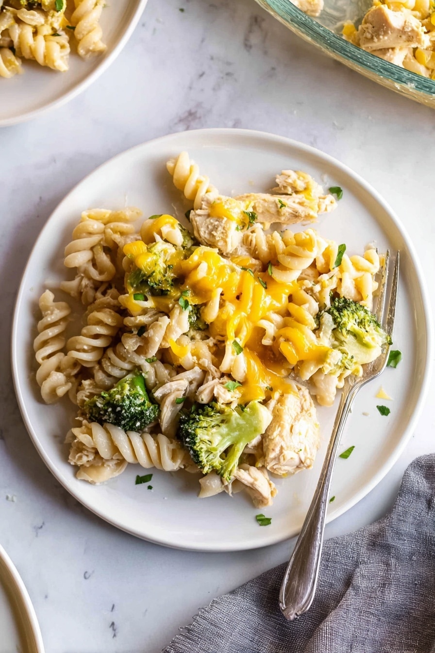 A white plate on a white marbled surface holds a serving of rotini pasta mixed with chunks of cooked chicken, bright green broccoli florets, and melted yellow cheese drizzled unevenly over the top. The pasta spirals are pale cream and slightly glossy, nestled closely with tender pieces of chicken and small green broccoli heads, all lightly sprinkled with small bits of fresh green herbs. A silver fork rests partially on the right edge of the plate, with its tines touching some pasta. There is a gray cloth napkin in the lower right corner and a partial view of another white plate and a glass baking dish in the background. Photo taken with an iphone --ar 2:3 --v 7 - Easy Chicken Noodle Casserole, chicken noodle casserole, creamy chicken pasta bake, family-friendly dinner recipes, simple casserole ideas