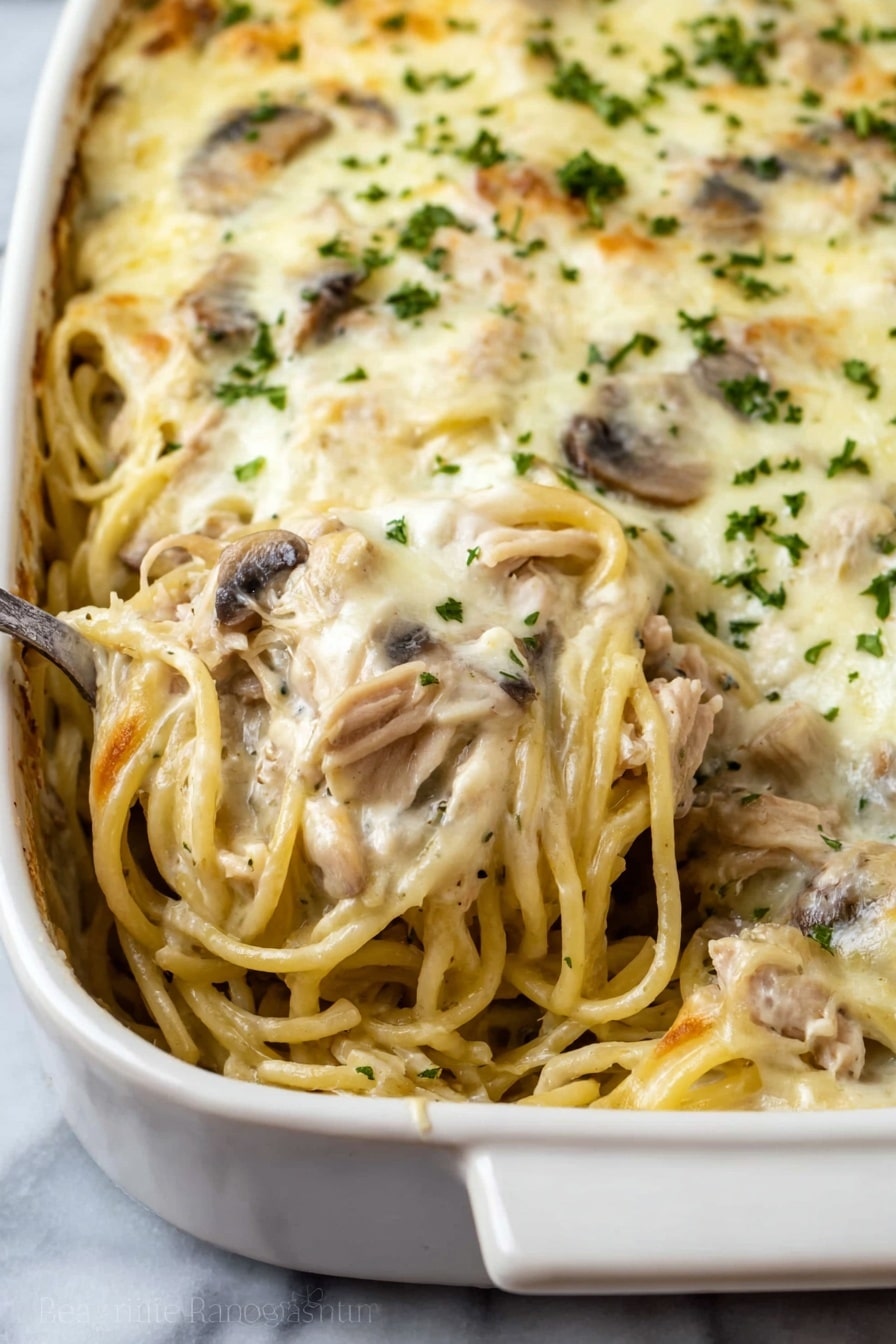 This dish shows a close-up view of a baked pasta casserole in a white ceramic dish. The casserole consists of creamy spaghetti mixed with pieces of light brown cooked meat and sliced mushrooms spread throughout. The top layer is covered with a smooth, melted white cheese sauce sprinkled with small bits of green herbs. A spoon is scooping some of the creamy pasta from the dish, showing the mixed layers of noodles, meat, and mushrooms inside. The dish is placed on a white marbled surface. photo taken with an iphone --ar 2:3 --v 7 - Creamy Chicken Tetrazzini Casserole, chicken tetrazzini with mushrooms, cheesy chicken casserole, easy chicken tetrazzini recipe, comforting chicken casserole