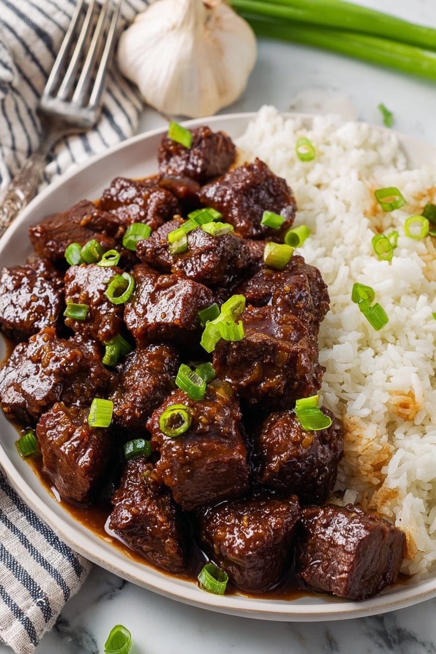 The image shows a white plate filled with two main layers: on the left and center, there are glossy, dark brown cubes of beef coated in a thick sauce, with chopped green onions sprinkled over the top, and on the right side, there is a layer of white rice with some sauce seeping into it. The plate is placed on a white marbled surface with a garlic bulb, a striped cloth, and a fork nearby. The focus is close, highlighting the texture of the tender beef and the fresh green garnish. Photo taken with an iphone --ar 2:3 --v 7 - Honey Garlic Steak Bites, Honey Garlic Steak Bites Recipe, Easy Steak Bites with Honey and Garlic, Juicy Steak Bites, Savory Honey Garlic Steak Bites
