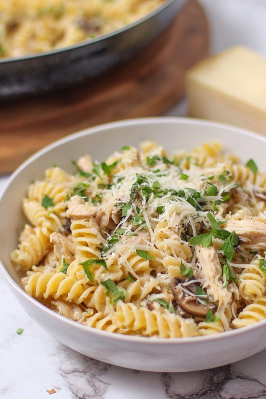 The image shows a white bowl filled with spiral pasta layered with light-colored shredded chicken and small bits of mushrooms. The top layer has finely grated cheese scattered evenly, which looks soft and slightly fluffy, and fresh green herb leaves spread all over, adding a fresh contrast. The background has a white marbled texture with a piece of cheese and a pan of more pasta slightly out of focus. The overall look is creamy and fresh, with natural light highlighting the details of the pasta and herbs. photo taken with an iphone --ar 2:3 --v 7 - Creamy Turkey Pasta, easy Thanksgiving leftovers dinner, comforting pasta with turkey, quick turkey pasta recipe, rich creamy pasta dish