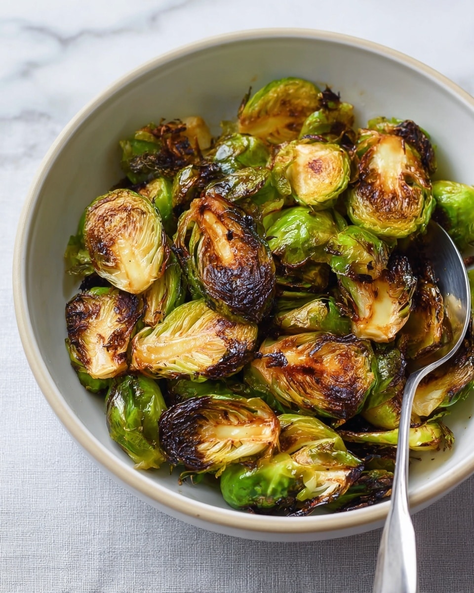 A white bowl filled with roasted Brussels sprouts, showing about two layers of vegetables. The Brussels sprouts are mostly green with many pieces having dark brown, slightly crisp edges. They have a shiny, slightly oily texture. Some loose, crispy, dark brown leaves are scattered among the whole and halved Brussels sprouts. A silver spoon rests inside the bowl on the right side. The bowl sits on a white marbled surface. Photo taken with an iphone --ar 4:5 --v 7