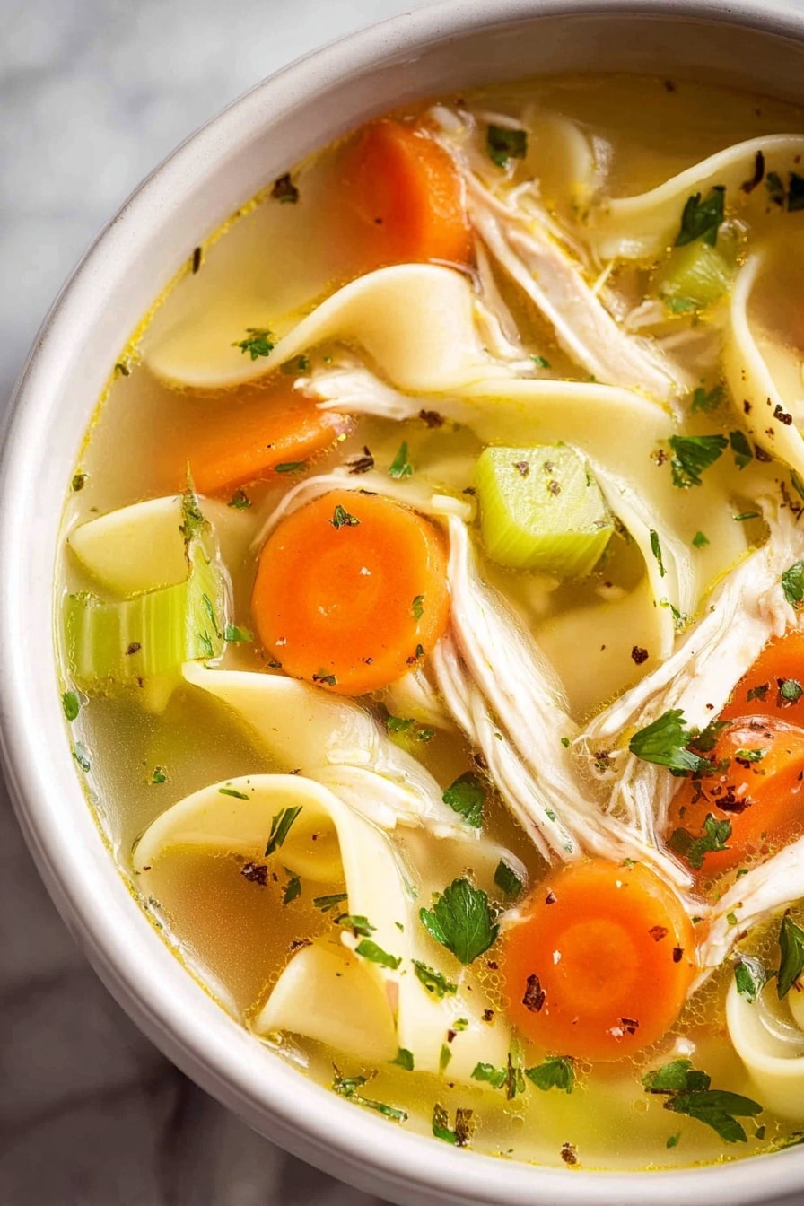 A white bowl filled with clear golden broth containing long, flat yellow noodles, small orange carrot cubes, and green celery pieces. Shredded light beige chicken is spread evenly throughout, with flecks of green herbs and black pepper for extra color. A single dark brown bay leaf is visible near the bottom, and a silver spoon is partially submerged in the soup. The bowl rests on a white marbled surface with a small white cup of chopped green herbs and another white cup with a white creamy substance in the background, photo taken with an iphone --ar 2:3 --v 7 - Homemade Chicken Noodle Soup from Scratch, hearty chicken noodle soup, homemade chicken soup recipe, from scratch chicken noodle soup, comforting chicken noodle soup