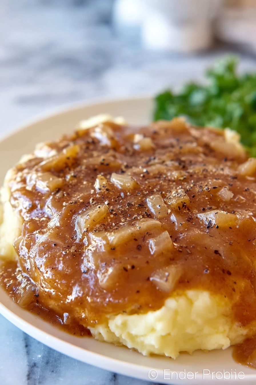 A white plate with mashed potatoes as the base layer, creamy and light yellow in color with a smooth texture, topped with a thick brown gravy that has small diced onion pieces spread evenly across the surface. The gravy covers a large piece of meat underneath, sprinkled with black pepper flakes. The background shows an out-of-focus white marbled surface and some green garnish in the back. Photo taken with an iphone --ar 2:3 --v 7 - Crock Pot Cube Steak, slow cooker cube steak recipe, easy crock pot steak, tender cube steak slow cooker, comforting slow cooker dinner