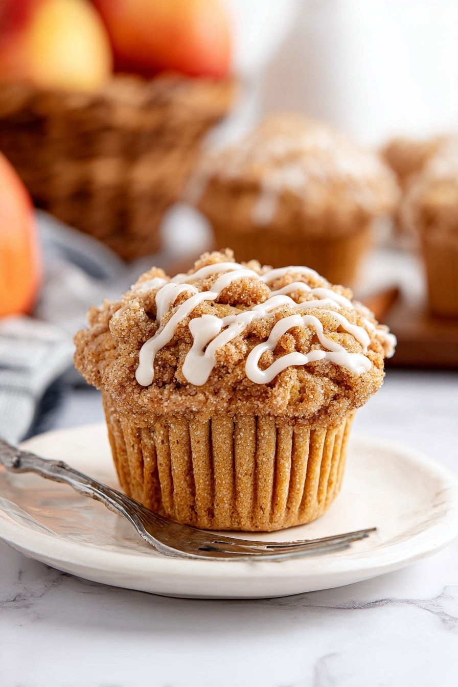 A single muffin sits centered on a white plate with a silver fork resting next to it. The muffin has a light brown, soft textured base with a crumbly streusel topping in a darker brown shade. White icing drizzle runs in thin lines over the streusel, adding contrast. The plate and muffin are placed on a white marbled surface. In the background, blurred muffins and a basket of apples add warm tones but stay out of focus. Photo taken with an iphone --ar 2:3 --v 7 - Cinnamon Apple Muffins with Crumble Topping and Glaze, fall breakfast muffins, spiced apple muffins, easy apple muffins recipe, cozy morning muffins