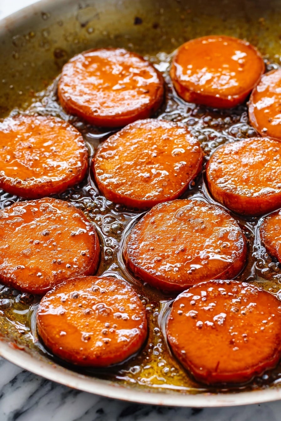 The image shows a white oval dish filled with thick, round slices of cooked sweet potatoes. The sweet potato slices are covered in a shiny, dark caramel-like sauce that makes them look glossy and soft. The slices are arranged closely together, slightly overlapping, and their orange color is rich and warm, enhanced by the dark syrup. The sauce pools around the edges of the slices, filling the bottom of the dish, and the texture looks smooth and sticky. The setting is on a white marbled surface. photo taken with an iphone --ar 2:3 --v 7 - Candied Sweet Potatoes with Bourbon Glaze, sweet potato side dish, bourbon glazed sweet potatoes, caramelized sweet potato recipe, festive holiday sweet potatoes