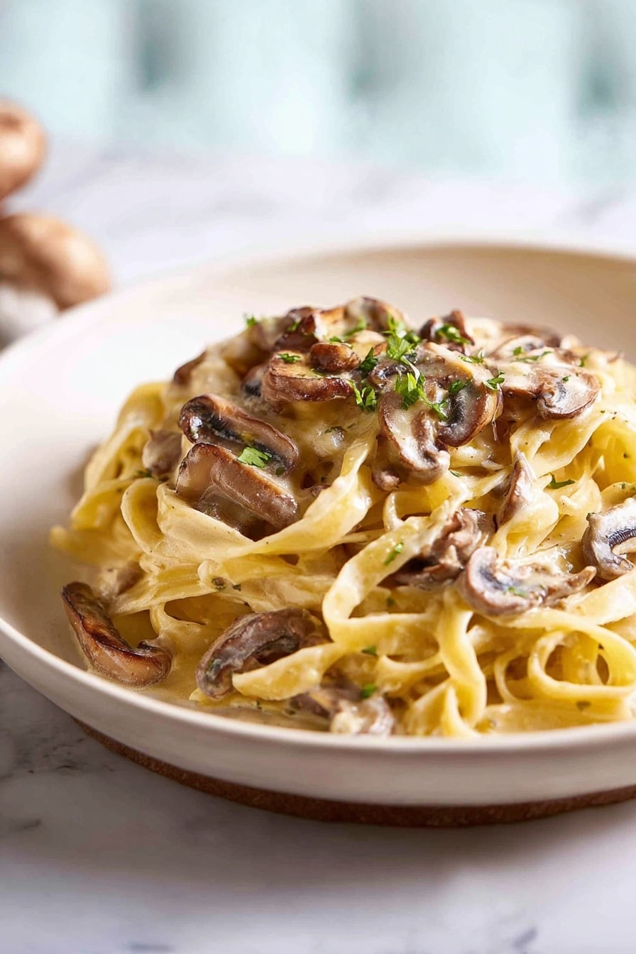 The image shows a mound of creamy fettuccine pasta on a plain white plate. The pasta is light yellow and looks soft and smooth, with thin sauce coating each strand. On top and mixed through are brown sliced mushrooms with a slight gloss, adding texture and earthy colors. Small green herb bits are sprinkled over the pasta, giving little pops of color. The plate sits on a white marbled surface, with a blurred background that is softly lit showing a light blue and white shade. Photo taken with an iphone --ar 2:3 --v 7 - Creamy Mushroom Pasta, mushroom pasta recipe, creamy mushroom pasta dinner, easy mushroom pasta, vegetarian creamy pasta