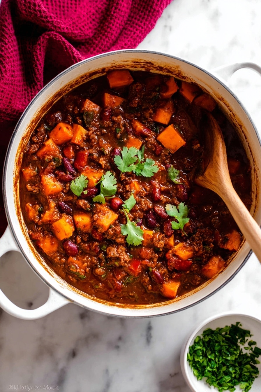 A white pot filled with thick stew that has three main layers: a dark brown sauce base, chunky orange sweet potato pieces, and visible red and brown beans mixed with ground meat; a few green cilantro leaves float on top near the center for color contrast. A wooden spoon is partially dipped on the right side, stirring the stew, showing the dense texture and rich sauce coating all ingredients. The pot rests on a white marbled surface with a dark red cloth draped in the upper left corner and a small white bowl filled with chopped green herbs placed near the bottom right. Photo taken with an iphone --ar 2:3 --v 7 - Vegetarian Pumpkin Chili, Best Pumpkin Chili, Easy Vegetarian Chili, Hearty Pumpkin Chili Recipe, Cozy Fall Chili