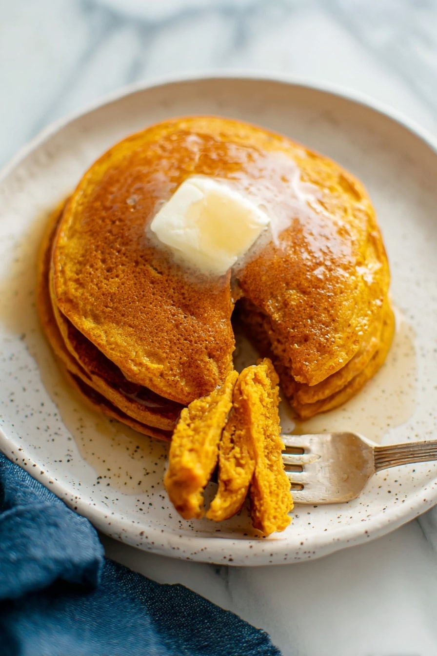 There is a white plate with three golden-brown pancakes stacked slightly unevenly with a clear textured top pancake showing tiny holes. A gold fork rests on the top edge of the plate. Next to the plate on the white marbled surface, there is a pile of more pancakes on another white plate that shows their soft, porous texture. A small white jug with dark syrup is near the plates, and a blue cloth is placed on the surface beside the plate. The photo taken with an iphone --ar 2:3 --v 7 - Whole Wheat Pumpkin Pancakes, healthy pumpkin pancake recipe, fall breakfast ideas, wholesome pumpkin pancakes, easy pumpkin pancake recipe