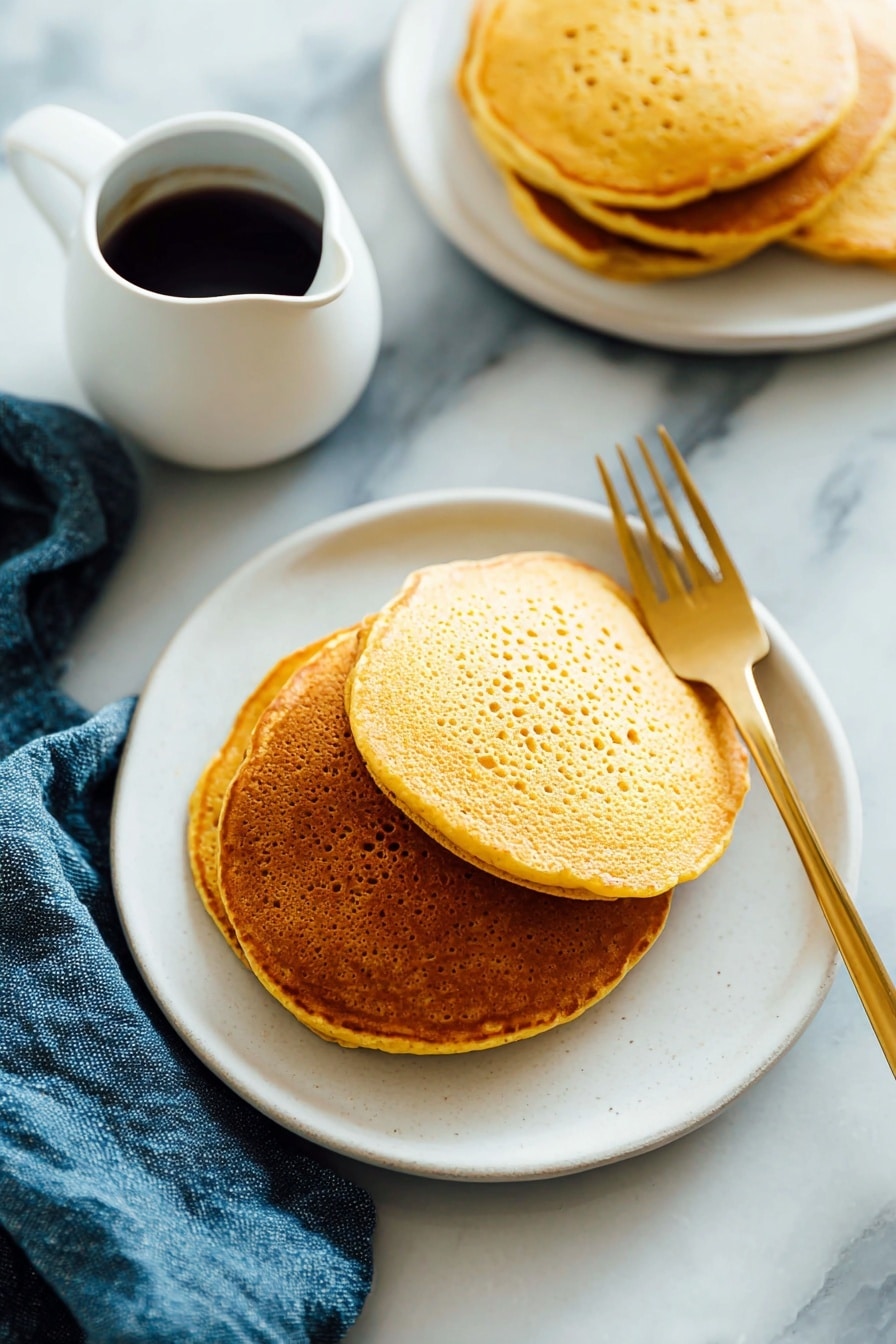 There is a stack of three golden-brown pancakes on a white plate. The pancakes have a slightly porous texture and are arranged with the largest at the bottom and the smallest on top. A light amber syrup is poured over the top pancake, dripping down the sides and pooling a little on the plate. A silver fork rests under the stack, on the left side, and the plate sits on a white marbled surface with a blue cloth nearby. The lighting is soft and natural, highlighting the warm tones of the pancakes and syrup. photo taken with an iphone --ar 2:3 --v 7 - Whole Wheat Pumpkin Pancakes, healthy pumpkin pancake recipe, fall breakfast ideas, wholesome pumpkin pancakes, easy pumpkin pancake recipe