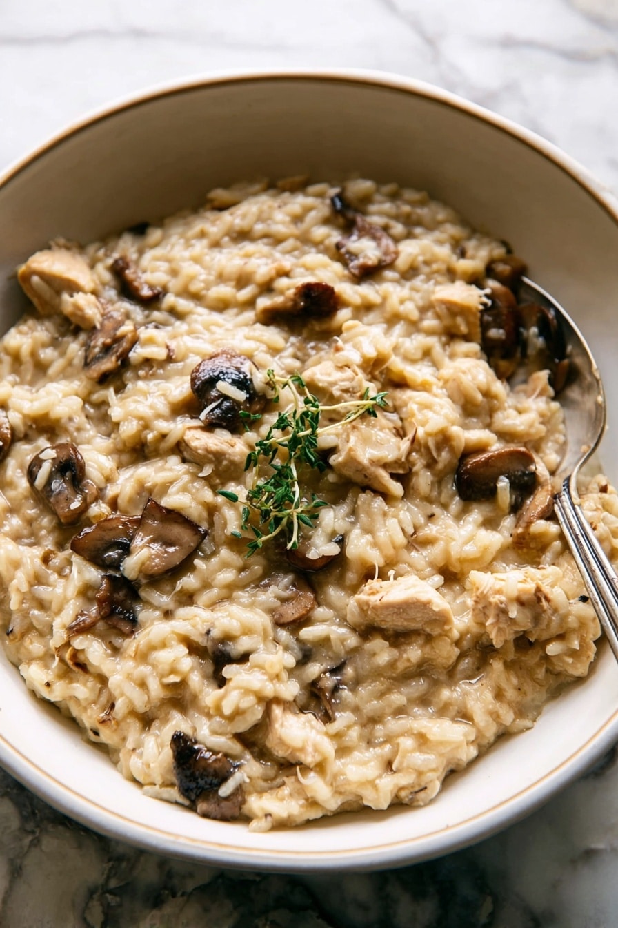A close-up of creamy risotto served in a white bowl with a pale green-blue inner glaze texture. The risotto has a light beige color with soft, plump rice grains and visible chunks of light brown chicken and dark brown mushrooms. A small green herb sprig rests on top as garnish. A silver fork sits on the right side partially stuck into the risotto. The background is a white marbled texture with a few out-of-focus green herb stems visible at the top. photo taken with an iphone --ar 2:3 --v 7 - Chicken Mushroom Risotto with Thyme, creamy chicken mushroom risotto, easy mushroom risotto recipe, comforting weeknight risotto, thyme flavored risotto