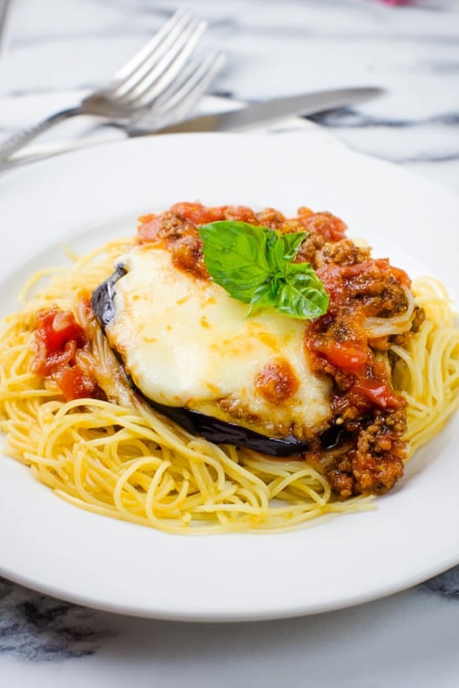 A white plate holds a dish with a bottom layer of thin yellow spaghetti noodles spread evenly. On top of the noodles are two thick slices of eggplant covered with a melted, light golden layer of cheese that looks soft and slightly browned in spots. A chunky meat and tomato sauce with visible small pieces of tomato and meat is spooned over the eggplant slices. A fresh green basil leaf is placed on top as garnish. The plate sits on a white marbled surface with a fork and knife just visible at the bottom left corner. Photo taken with an iphone --ar 2:3 --v 7 - Eggplant Parmesan Spaghetti with Meat Sauce, Eggplant Parmesan spaghetti, hearty Italian pasta recipes, crispy eggplant pasta dish, meat sauce spaghetti dinner