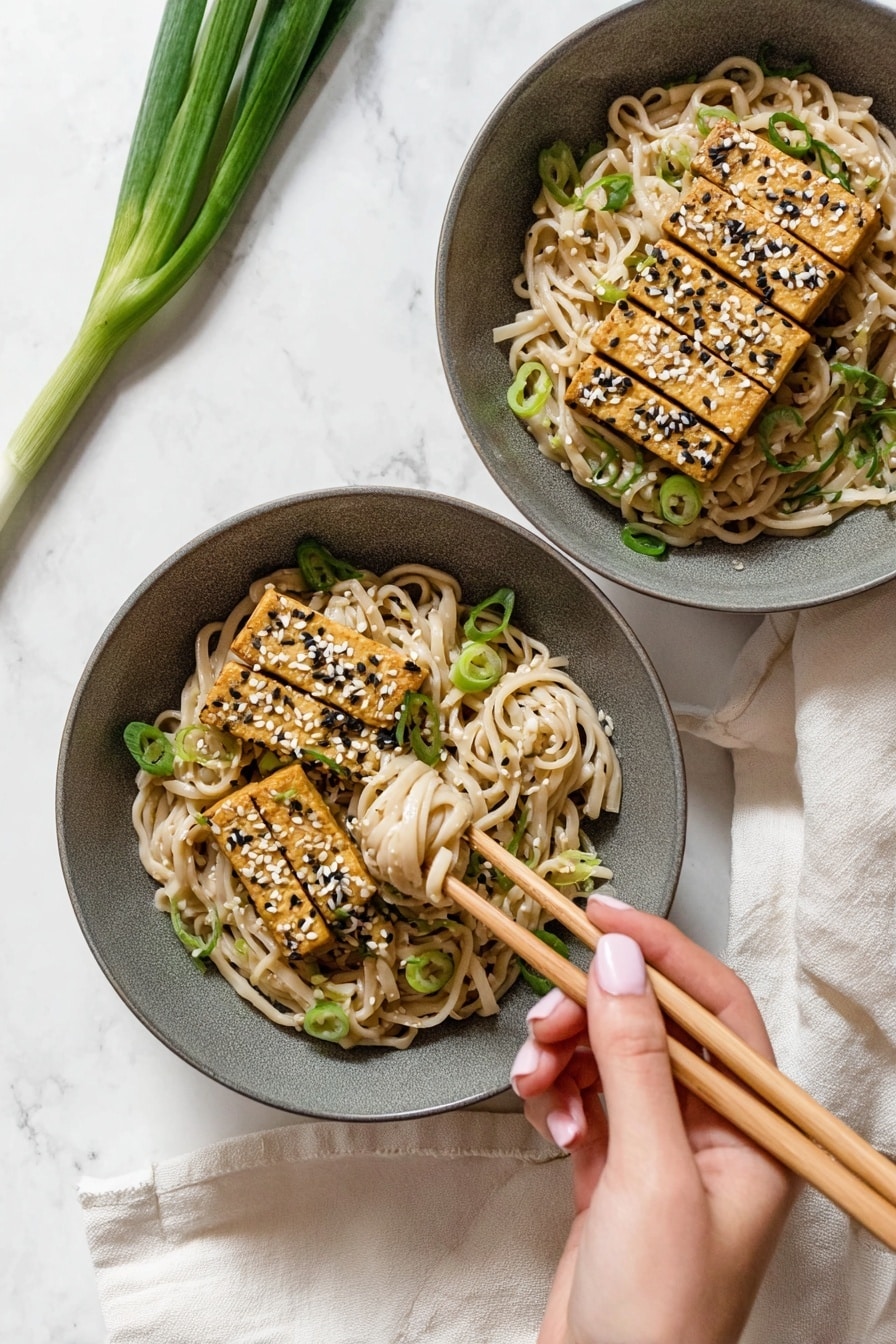 A round white bowl filled with creamy light beige noodles mixed with small green pieces, topped with four rectangular golden brown tofu slices sprinkled with white and black sesame seeds and chopped green onions. The noodles have a smooth texture, and the tofu pieces are placed side by side in the center. The bowl is set on a white marbled surface with two whole green onions placed above it and a pair of wooden chopsticks resting on a beige cloth to the right. Photo taken with an iphone --ar 2:3 --v 7 - Easy Peanut Udon Noodles, peanut udon noodles, quick udon recipes, Asian noodle dishes, flavorful udon stir-fry