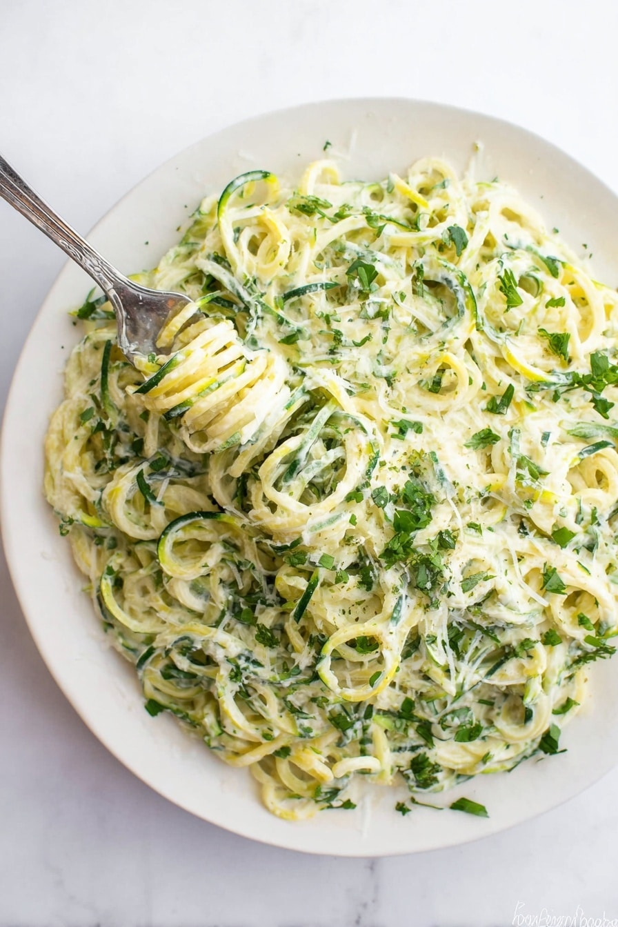 A white plate filled with a creamy dish made of spiral-shaped green and light yellow vegetable noodles, mixed with a white creamy sauce that coats the noodles evenly. There are small green herb leaves sprinkled throughout the dish, adding a fresh look. A silver fork is twirling some of the noodles in the top left area of the plate. The plate rests on a white marbled surface. photo taken with an iphone --ar 2:3 --v 7 - Creamy Zucchini Alfredo, healthy zucchini Alfredo, easy zucchini noodle recipe, light creamy pasta, quick dinner with zucchini