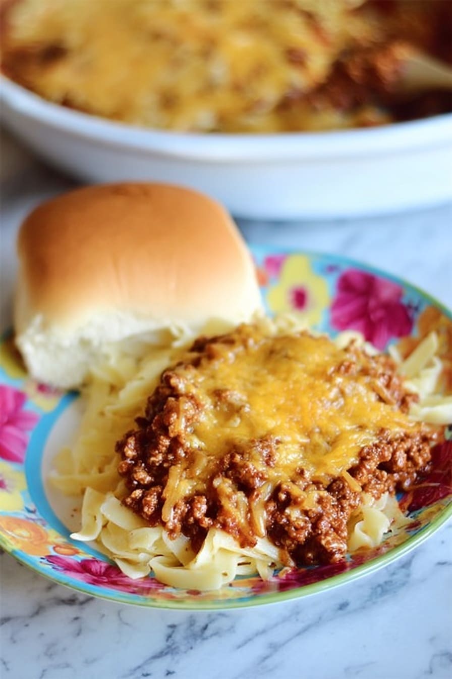 The dish shows a serving of pasta on a white plate with a colorful flower design in pink, red, and yellow. The pasta forms the bottom layer with visible white and slightly curved noodles. On top of the pasta, there is a thick layer of cooked ground meat in dark brown color mixed with reddish-brown sauce. The top layer is melted orange-brown cheese covering most of the meat. Next to the pasta is a soft, golden-brown bread roll with a smooth surface and light cracks. The background shows a white round dish filled with the same pasta, meat, and cheese mix. The whole scene is set on a white marbled surface. Photo taken with an iphone --ar 2:3 --v 7 - Sour Cream Noodle Bake with Ground Beef, hearty ground beef casserole, creamy beef noodle bake, easy comfort food recipes, family-friendly dinner ideas