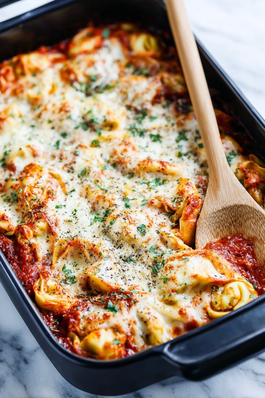 A black baking dish holds a layered tortellini pasta bake with a rich tomato sauce. The top layer is melted white cheese, bubbly and slightly browned in spots, mixed with twisted yellow tortellini filled pasta visible underneath. The sauce is deep red with bits of herbs and small chunks of meat mixed in. Scattered green herb leaves add color contrast across the top. The dish rests on a white marbled surface with a folded striped cloth nearby and a wooden-handled black spoon on the right edge of the frame, creating a cozy kitchen feel. photo taken with an iphone --ar 2:3 --v 7 - Cheesy Baked Tortellini Casserole, cheese tortellini casserole, baked tortellini recipes, meat tortellini bake, easy comfort food dinner