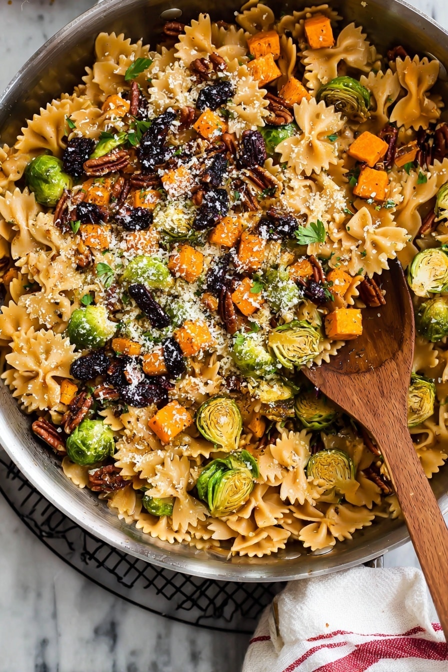 A large silver pan filled with a colorful pasta dish featuring three layers: the bottom layer has light brown farfalle pasta with smooth, slightly shiny texture; the middle layer contains bright orange small cubed roasted vegetables and sliced green brussels sprouts, some with charred marks, scattered evenly; the top layer is sprinkled with chopped dark brown pecans, black dried fruit pieces, small green herb leaves, and a light dusting of finely grated white cheese. A wooden spoon with a rounded bowl sits inside the pan. The pan rests on a black wire rack with a white cloth with red stripes on the side, all set on a white marbled surface. Photo taken with an iphone --ar 2:3 --v 7 - Brussels Sprouts Pasta with Brown Butter, Brussels Sprouts Pasta, brown butter pasta, healthy Brussels sprouts recipes, cozy vegetarian pasta