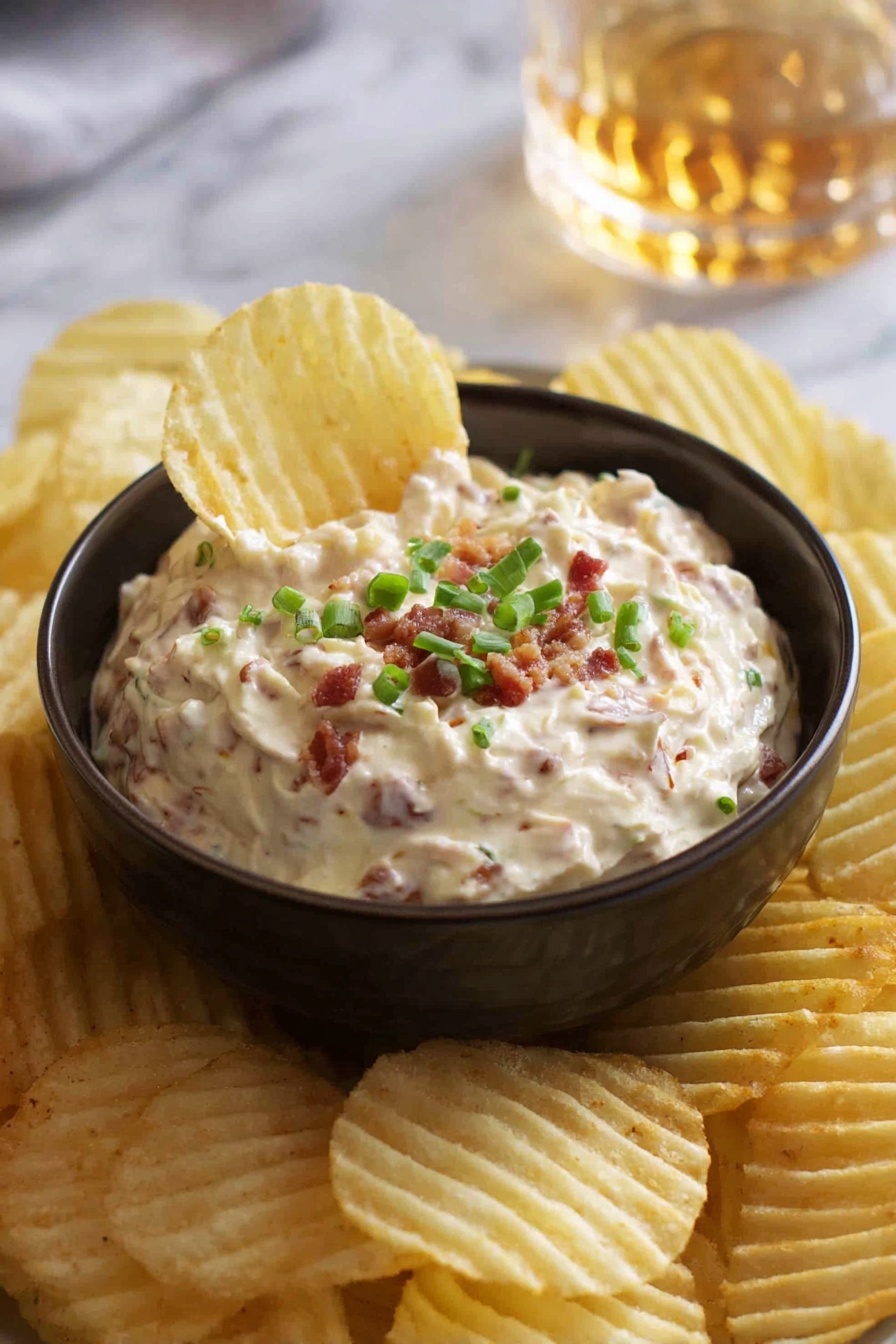 A small dark bowl filled with creamy dip that has small pieces of bacon mixed in, topped with chopped green onions. The bowl sits on a white marbled surface and is surrounded by many ridged potato chips, one chip is dipped into the bowl showing the texture of the dip. In the background, there is a blurred glass with a golden liquid. Photo taken with an iphone --ar 2:3 --v 7 - French Onion Dip, homemade French onion dip, easy French onion dip, creamy onion dip, savory onion dip