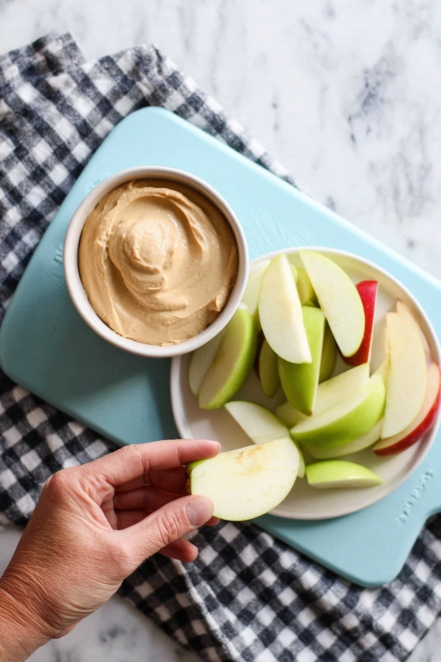 A woman's hand is holding a pale green apple slice dipped in a small bowl of light brown creamy dip. Next to it, on a white plate, there is a pile of apple slices in green and red colors. Both the bowl and plate are placed on a light blue cutting board, which rests on a black and white checkered cloth on a white marbled surface. The scene is brightly lit, showing clear textures of the creamy dip and crisp apple slices. photo taken with an iphone --ar 2:3 --v 7 - Easy Caramel Apple Dip, caramel apple dip, fall dip recipes, simple apple dip, quick party snacks