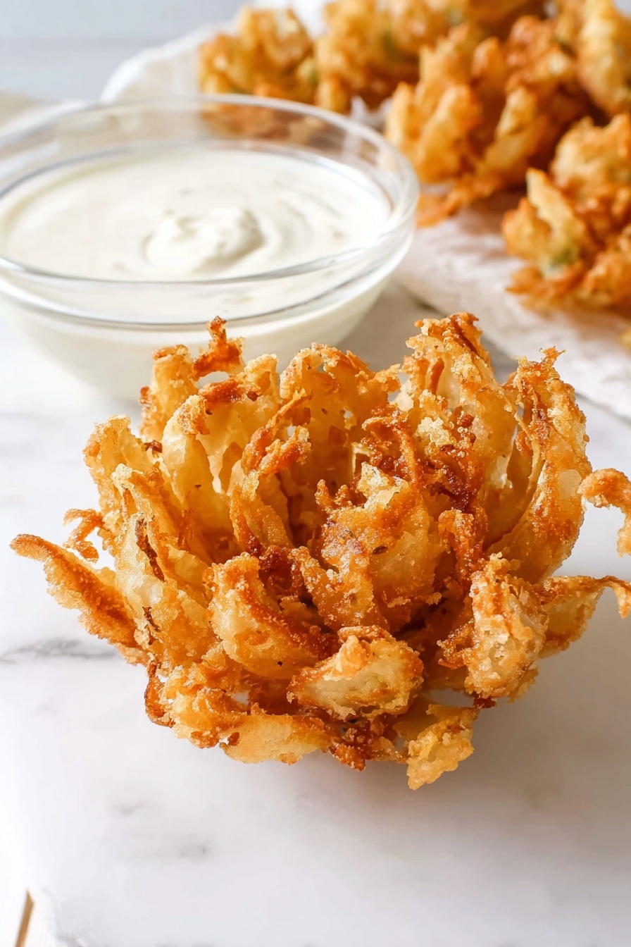 A close-up image of a golden-brown, crispy fried onion blossom placed on a white marbled surface. The onion blossom is fully opened like a flower with many uneven, crunchy petals showing a textured, fried batter coating. Next to it is a small clear glass bowl filled with smooth, creamy white dipping sauce. In the background, some more fried onion blossoms are slightly blurred. The scene is bright and simple with clean white tones and a soft shadow under the food. photo taken with an iphone --ar 2:3 --v 7 - Homemade Blooming Onion with Dip,Blooming Onion recipe, Crispy onion appetizer, Steakhouse-style onion petals, Easy party appetizer with dip