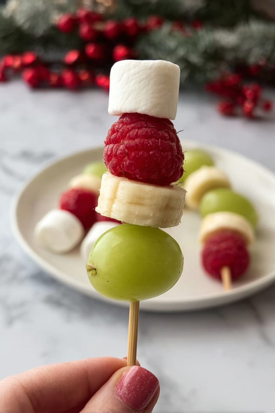 A small skewer held by a woman's hand shows four layers: at the bottom, a smooth, green grape; above it, a round, pale yellow banana slice; next a bright red raspberry with visible bumpy texture; and on top, a small white marshmallow shaped like a cube. In the background, more similar skewers rest on a white plate on a white marbled surface with some red berries. The overall look is fresh, colorful, and festive. photo taken with an iphone --ar 2:3 --v 7 - Grinch Fruit Kabobs, festive fruit kabobs, holiday fruit skewers, Christmas party snacks, easy holiday recipes
