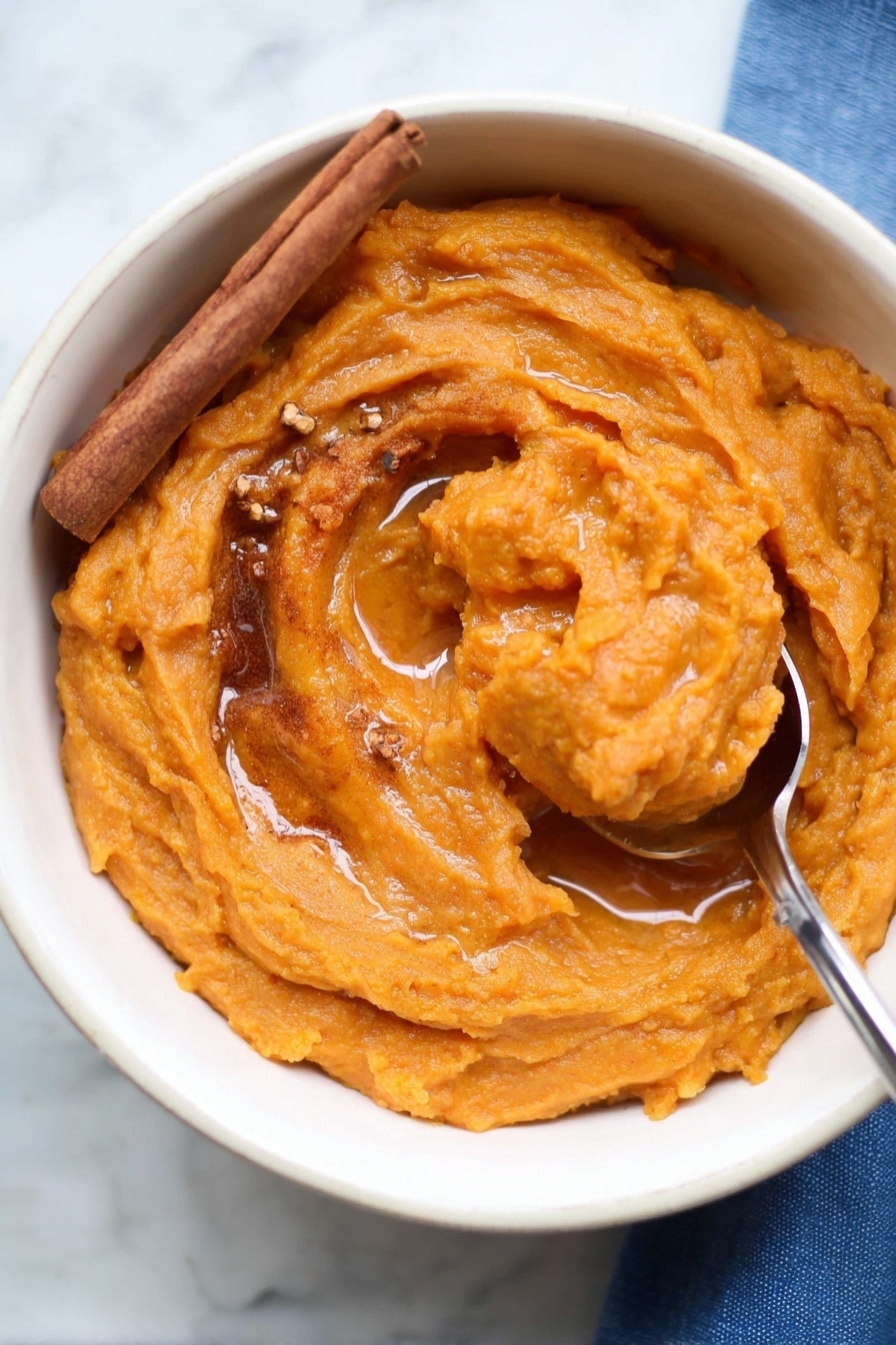 The image shows a white bowl filled with thick, smooth orange sweet potato mash. The mash has swirled texture with a shiny surface, topped with a light drizzle of syrup pooling slightly in the center. A cinnamon stick rests on the left side of the mash, and small bits of brown spices are sprinkled on top. A metal spoon is scooping up part of the mash on the right side, with the spoon handle extending out of the bowl. The bowl sits on a white marbled surface with a bit of a blue cloth visible in the bottom right corner. Photo taken with an iphone --ar 2:3 --v 7 - Brown Butter Mashed Sweet Potatoes, mashed sweet potato recipes, caramelized sweet potatoes, cozy side dishes, holiday vegetable sides