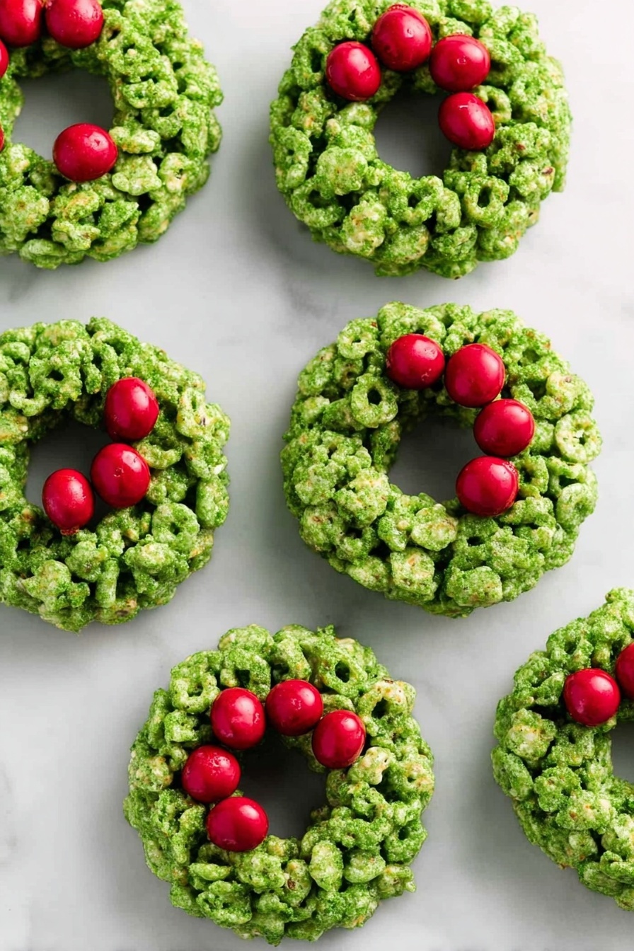 The image shows seven small green wreath-shaped treats placed on a white marbled surface. Each wreath is made from clusters of green cereal pieces stuck together, forming a thick ring with a hollow middle. Bright red round candies are grouped in threes on the top right side of each wreath, resembling berries on a holiday decoration. The green cereal has a rough texture with some visible cornflakes, and the red candies look smooth and shiny. The overall look is festive and colorful, with the wreaths evenly spaced across the surface. photo taken with an iphone --ar 2:3 --v 7 - Green Marshmallow Cornflake Wreaths, festive holiday treats, easy Christmas snacks, kid-friendly holiday recipes, colorful holiday wreaths