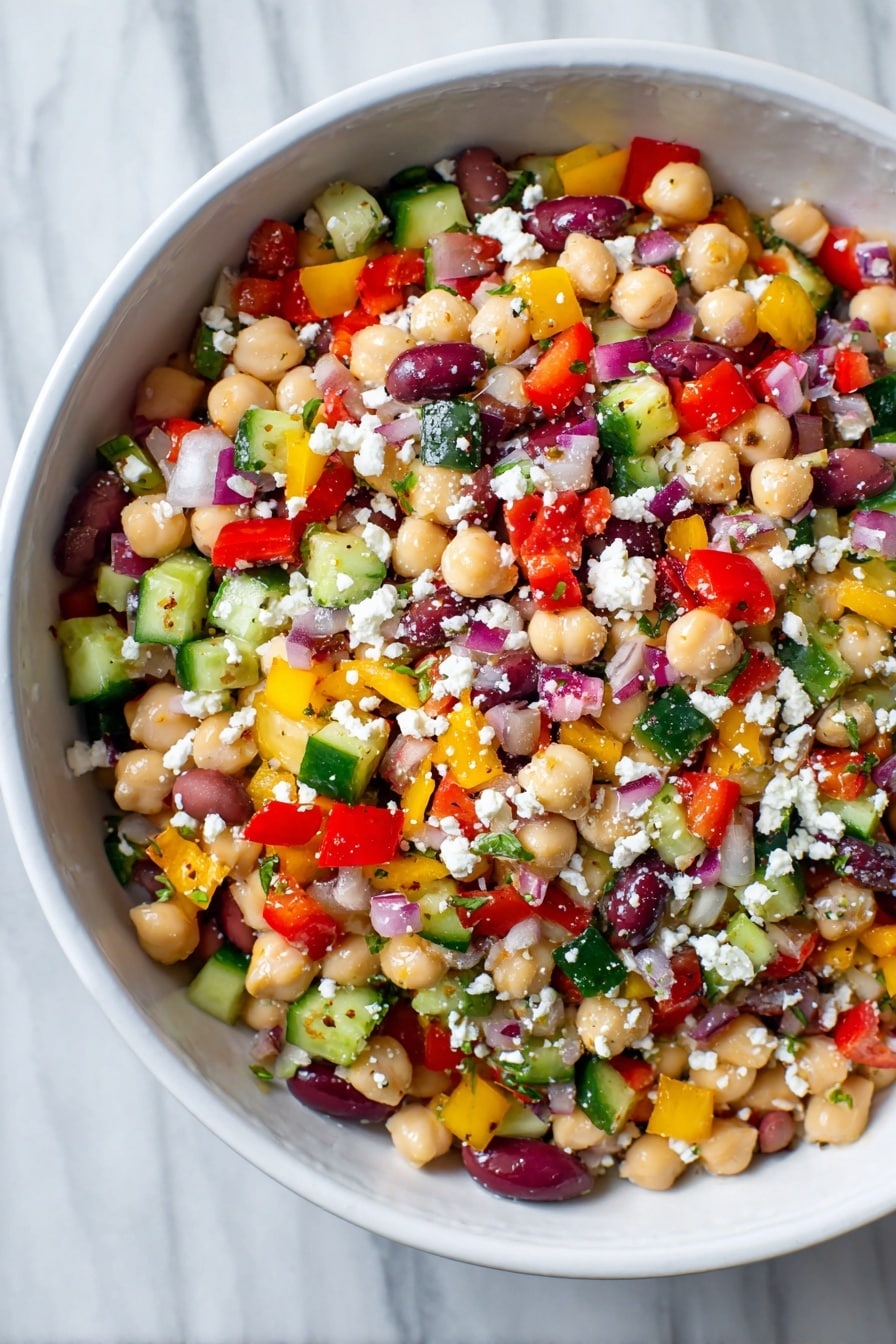 A white bowl filled with a colorful bean salad showing multiple layers of small, chopped vegetables and legumes. The base layer is made of light beige chickpeas and white beans mixed with a scattering of deep purple olive slices. On top, there is a thick mix of small diced bright red bell peppers, yellow bell peppers, green cucumbers, and red onions, creating a vibrant patchwork of colors. White crumbled cheese is sprinkled evenly over the salad, adding a soft, crumbly texture. The surface of the salad looks fresh, moist, and lightly coated in dressing. The bowl rests on a white marbled texture. Photo taken with an iphone --ar 2:3 --v 7 - Healthy Bean Salad with Feta, healthy bean salad, bean and feta salad, nutritious bean salad recipe, quick vegan bean salad
