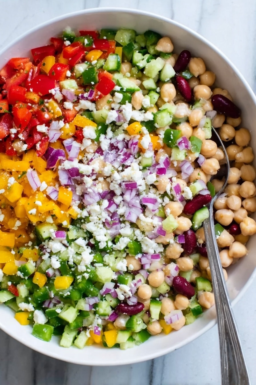 The image shows a white bowl filled with a colorful salad made of many small chopped vegetables and beans. There are five visible layers: at the bottom is a mix of small round chickpeas and white beans with a creamy texture, above that are chopped bright red tomatoes and diced yellow peppers spread evenly, next is a layer of finely chopped green celery and cucumber giving a fresh look, on top of that are small pieces of purple onions scattered across, and sprinkled all over are tiny white crumbles of cheese. A silver spoon is resting on the left side inside the bowl. The bowl sits on a white marbled surface. Photo taken with an iphone --ar 2:3 --v 7 - Healthy Bean Salad with Feta, healthy bean salad, bean and feta salad, nutritious bean salad recipe, quick vegan bean salad