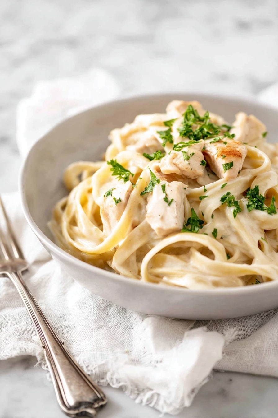A white bowl holds creamy fettuccine pasta topped with pieces of light golden cooked chicken. The pasta is thick and pale, coated with a smooth white sauce. There are small green bits of parsley sprinkled on top, adding a fresh touch. The bowl sits on a soft white cloth, placed on a white marbled surface. Nearby, a silver fork rests on the cloth. photo taken with an iphone --ar 2:3 --v 7 - One Pot Chicken Alfredo Pasta, One Pot Alfredo, Easy Chicken Alfredo, Creamy Chicken Pasta, Quick Alfredo Dinner