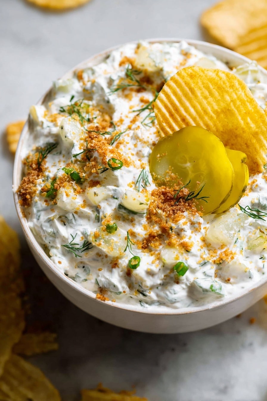 A white bowl filled with a creamy white dip mixed with small green herb pieces and light yellow chunks, topped with bright yellow pickle slices near the center. The dip is sprinkled with light brown crumbs and small green onion pieces, with a few small green herb sprigs scattered on top. A ridged golden potato chip is dipped into the creamy mixture near the edge of the bowl. The bowl sits on a white marbled surface with some crumbs and chips around it. photo taken with an iphone --ar 2:3 --v 7 - Creamy Ranch Pickle Dip, ranch dill pickle dip, easy pickle dip recipe, party snack dip, creamy appetizer with pickles