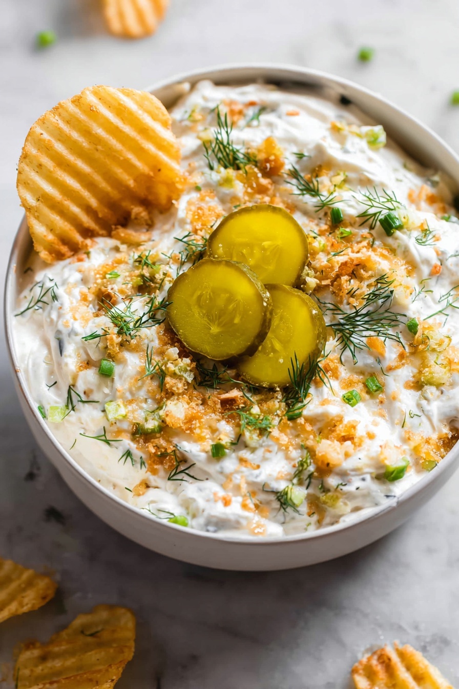 The image shows a grey bowl filled with a creamy white dip that is topped with several layers: small green chopped herbs, light brown crunchy bits, and three thick slices of pickles with a shiny, oily surface in the center. The dip looks thick and textured, with the crunchy bits scattered evenly on top. A woman’s hand is dipping a ridged, yellow chip into the creamy white mixture on one side. The bowl sits on a white marbled surface with some chips placed around it. photo taken with an iphone --ar 2:3 --v 7 - Creamy Ranch Pickle Dip, ranch dill pickle dip, easy pickle dip recipe, party snack dip, creamy appetizer with pickles