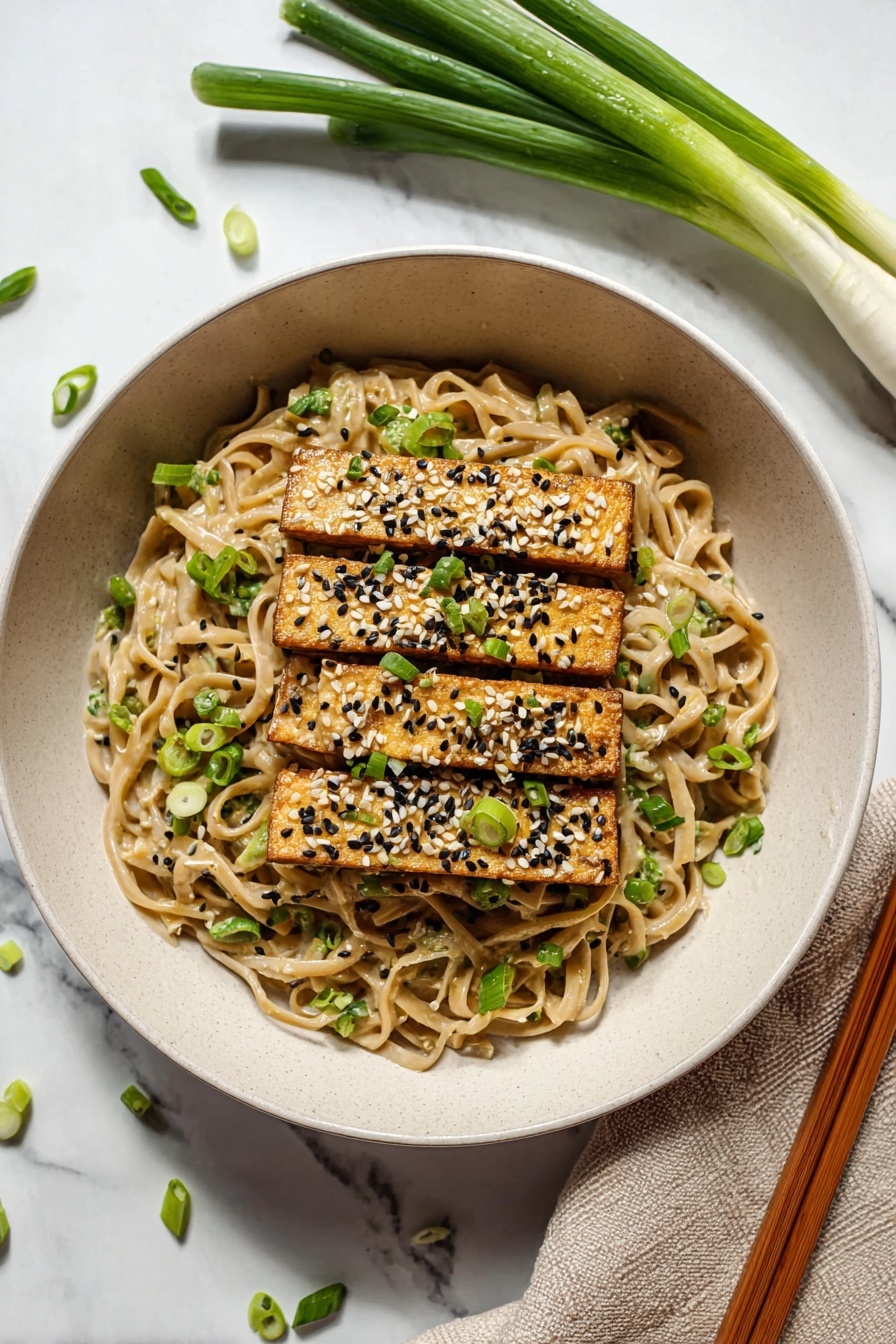 The image shows a close-up of a bowl with three layers. The bottom layer is thick, light tan noodles with a creamy sauce, speckled with tiny black and white seeds. On top of the noodles, there are golden-brown rectangular tofu pieces sprinkled with white and black sesame seeds and chopped green onions. At the top, a pair of wooden chopsticks held by a woman's hand lifts some noodles, with a few green onion slices stuck to the chopsticks. The bowl is white, and the background surface has a white marbled texture. photo taken with an iphone --ar 2:3 --v 7 - Easy Peanut Udon Noodles, peanut udon noodles, quick udon recipes, Asian noodle dishes, flavorful udon stir-fry