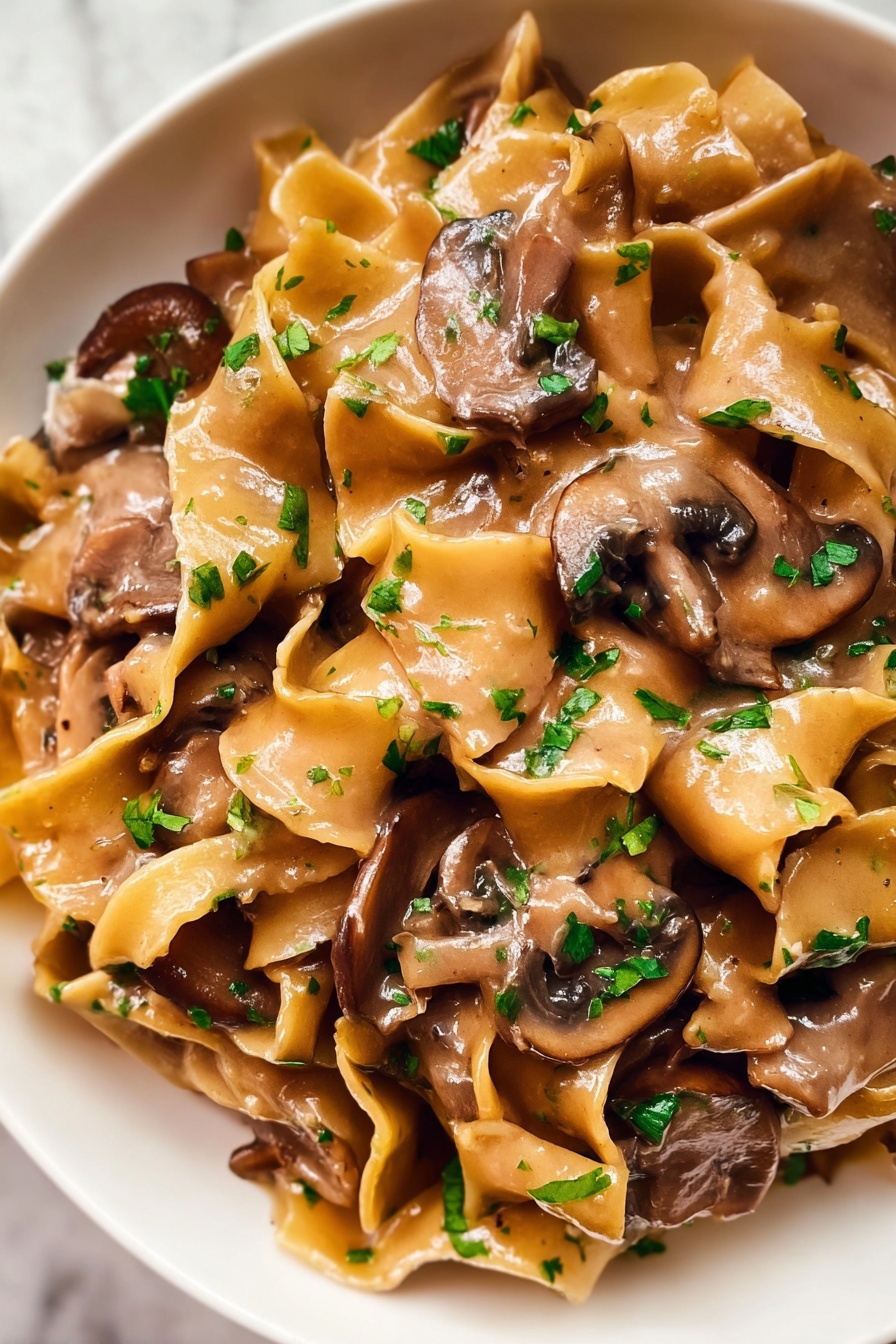 The image shows a close-up of a creamy pasta dish with wide, flat egg noodles mixed with thick slices of brown mushrooms. The noodles are coated in a smooth, light brown sauce that looks rich and glossy. Small pieces of chopped green herbs are sprinkled over the top, adding a fresh touch to the dish. The textures of the soft noodles and tender mushrooms blend together, showing a warm and comforting meal. The dish is served in a white bowl placed on a white marbled surface. photo taken with an iphone --ar 2:3 --v 7 - Creamy Vegetarian Mushroom Stroganoff, vegetarian mushroom steak, quick mushroom dinner, vegan mushroom sauce, easy vegetarian mushroom recipes