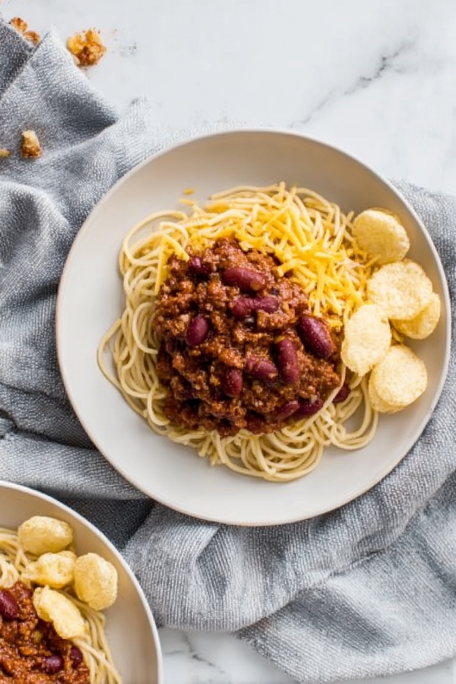 The image shows a white plate with three main layers. The bottom layer is a pile of light brown spaghetti noodles arranged in a loose small mound. On top of the noodles, there is a thick layer of dark reddish-brown chili with visible kidney beans and a chunky texture. To one side of the noodles and chili, there is a small group of round, pale beige crunchy chips neatly placed. The noodles and chips sit on a white marbled surface with a soft grey cloth draped around the plate. In the corner of the photo, part of another similar plate with the same food is visible. Photo taken with an iphone --ar 2:3 --v 7 - Cincinnati Chili Spaghetti, Cincinnati Chili, Chili Spaghetti Recipe, Comfort Food Recipes, Easy Cincinnati Chili