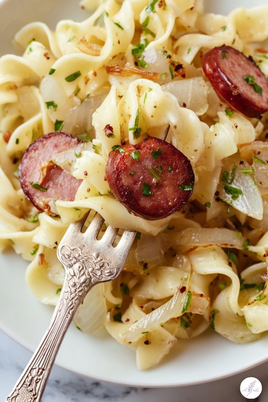 A close-up view of a white plate filled with twisted egg noodles that are light yellow with a soft texture, mixed with small pieces of cooked onions that are translucent white with a slight brown edge, and slices of browned sausage that are dark red with a grilled surface and visible herbs. The noodles and ingredients are sprinkled with chopped green herbs and black pepper. A vintage silver fork with floral engravings is lifting some noodles and one slice of sausage. The background is a white marbled surface. Photo taken with an iphone --ar 2:3 --v 7 - Easy Kielbasa and Cabbage Haluski, kielbasa and cabbage stir-fry, simple Polish haluski, hearty sausage and cabbage dish, quick comfort food recipes